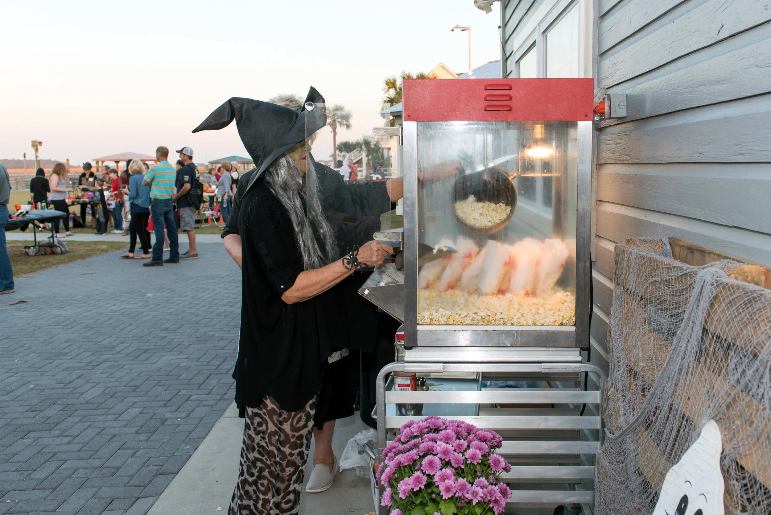 Person dressed as witch, scooping popcorn from machine at Halloween festival.