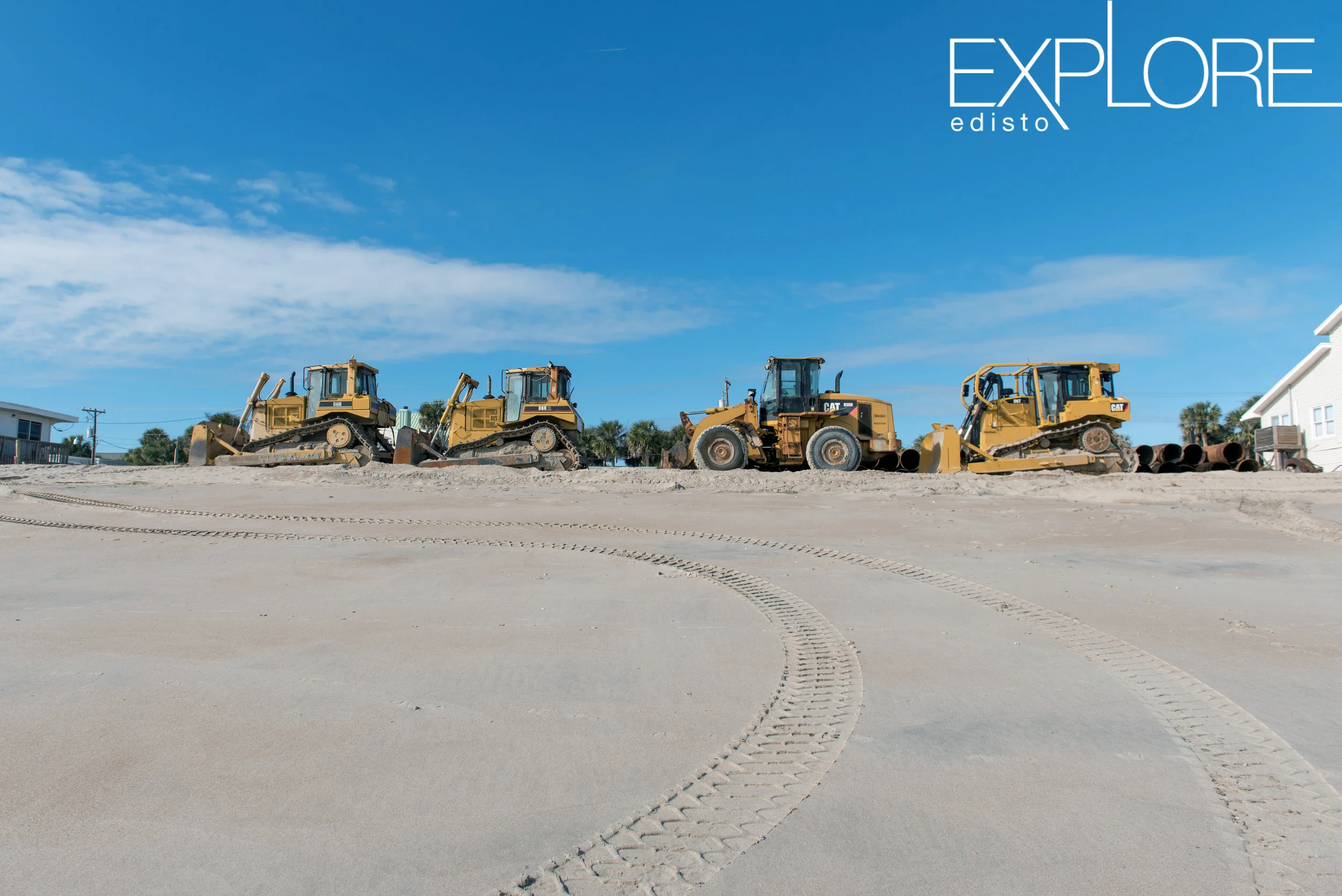 Four tractors lined up on the beach in front of blue sky.