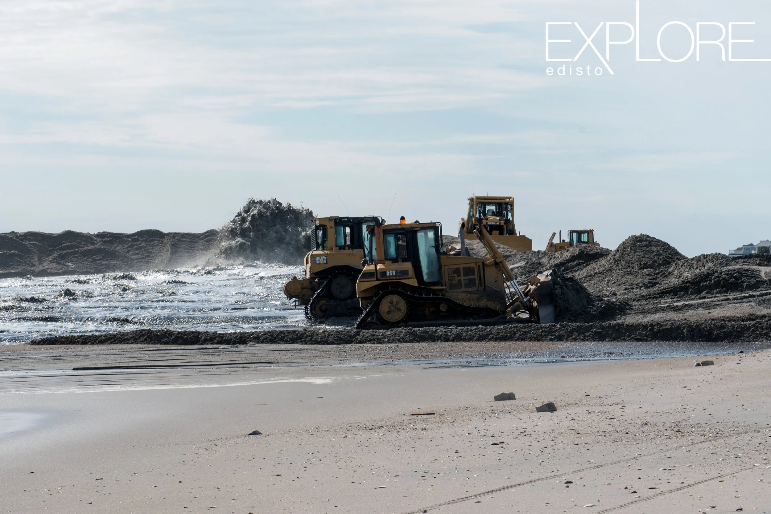 Tractors pushing sand onto the beach while it is being blasted to the shore from a pipe in the distance.