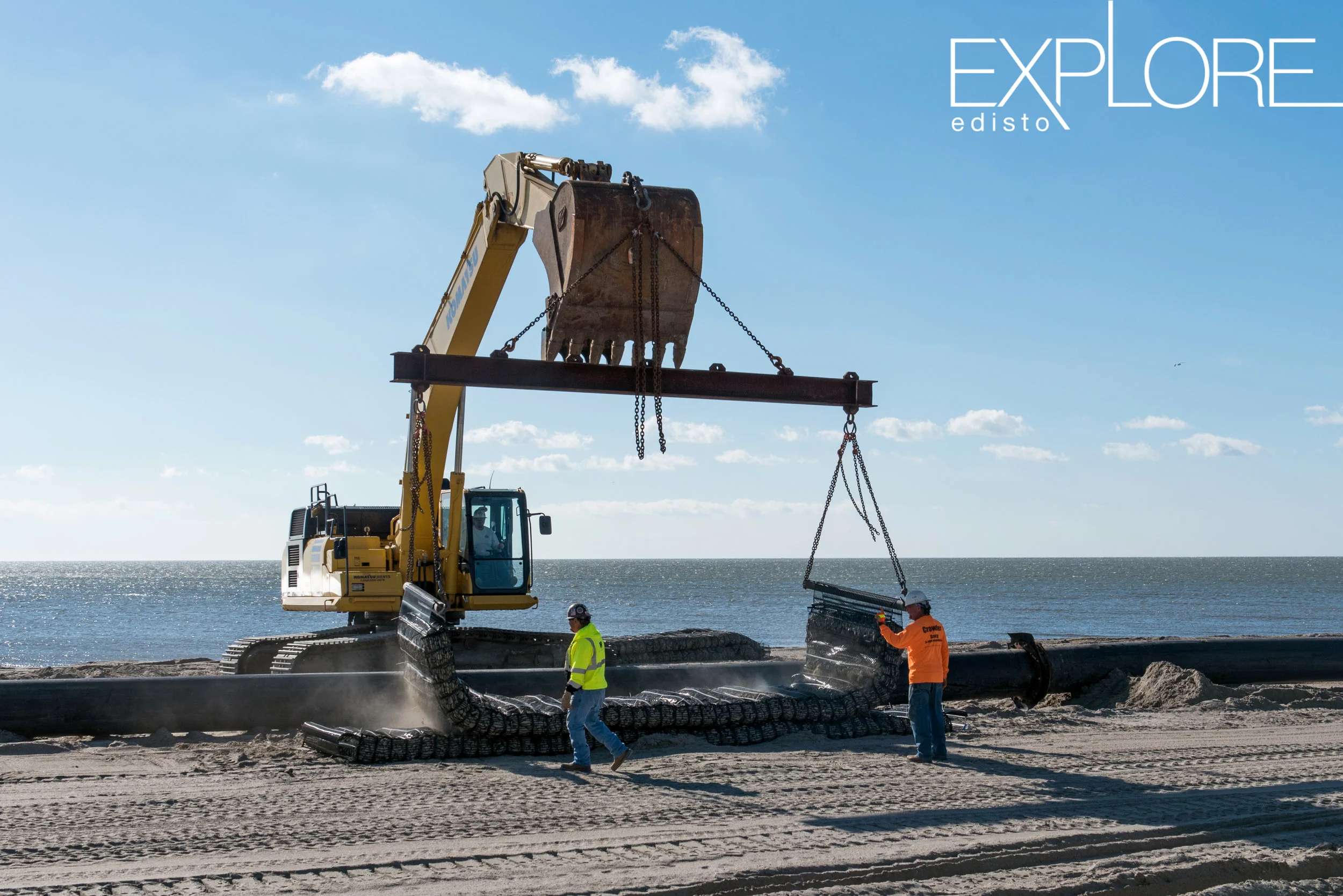 Large crane on the beach moving rocks with workers in hard hats and neon yellow and green shirts.