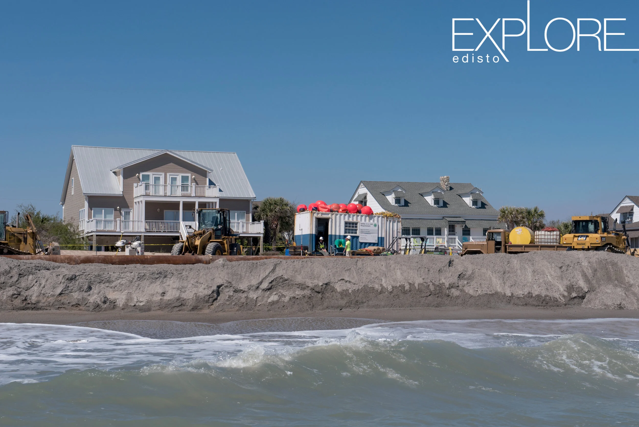 View of beach renourishment from the ocean. Container and tractors on beach with houses in background.