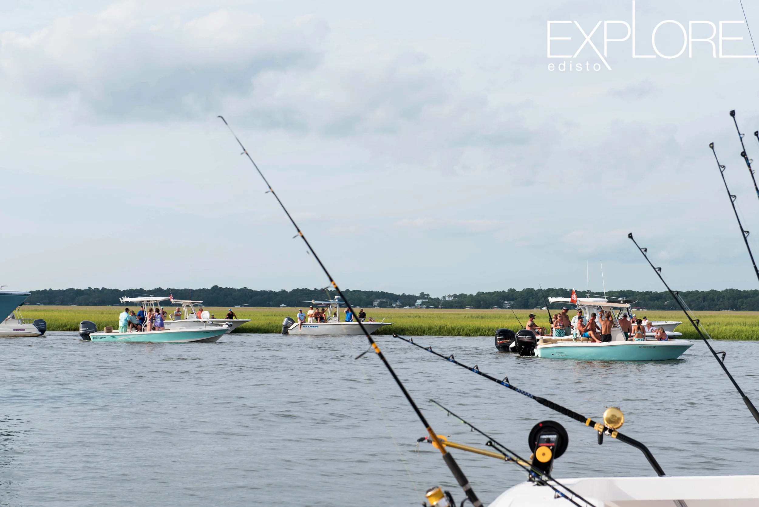 Fishing poles in foreground with several boats floating in creek in background.