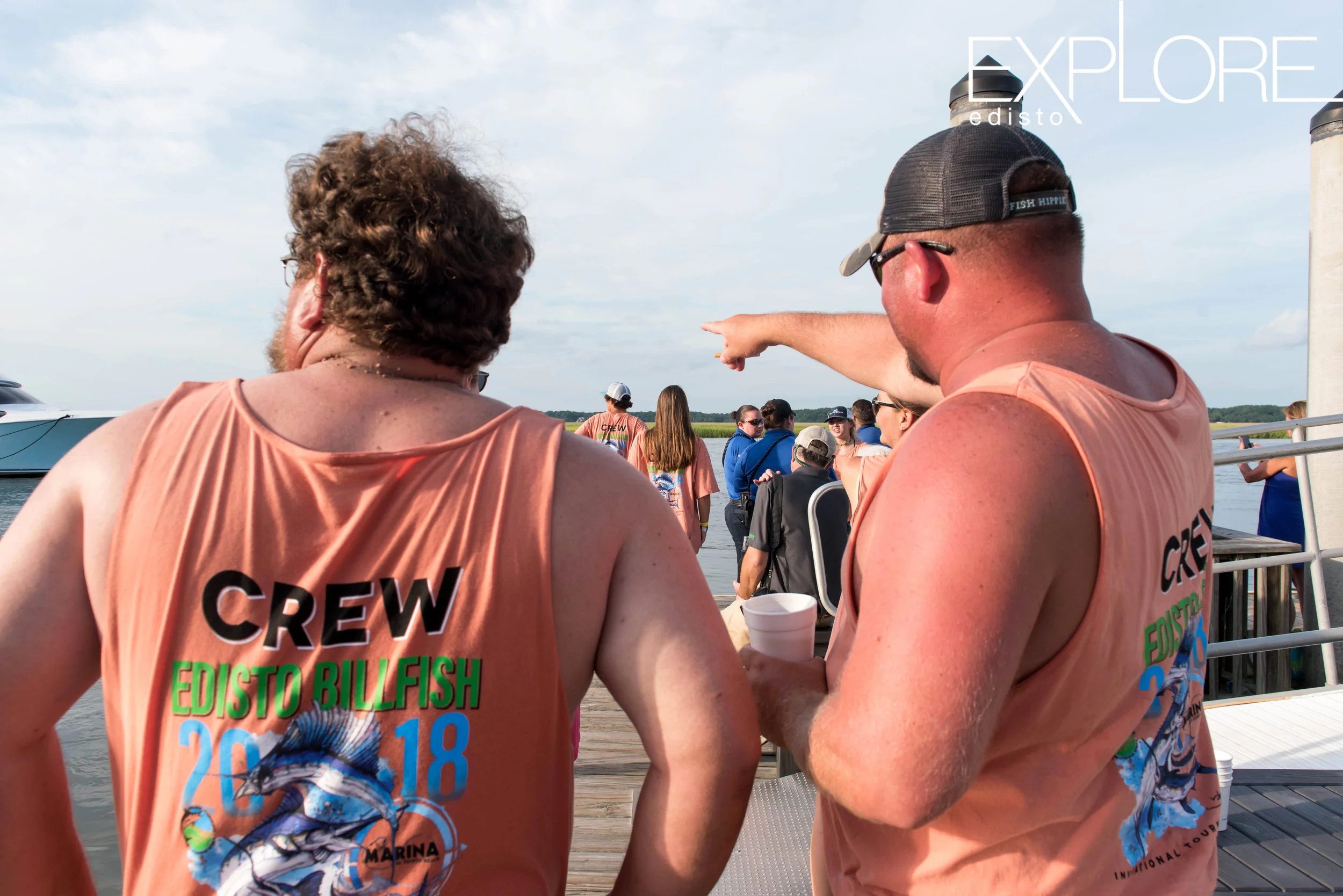 Men in orange "crew" tank tops look out in distance while one of them points.