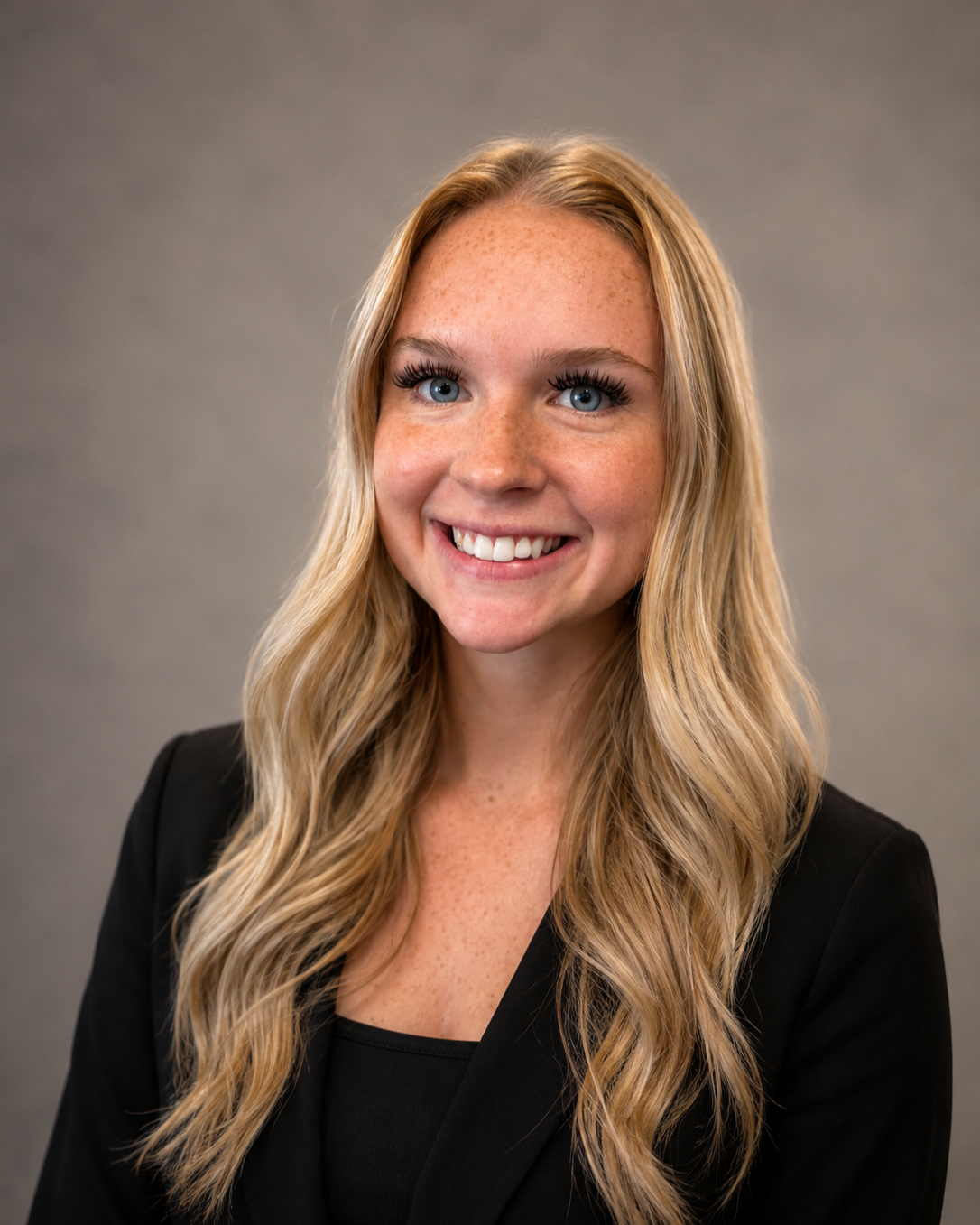 A young woman with long wavy blonde hair, blue eyes, and freckles smiling in a black blazer against a plain gray background.