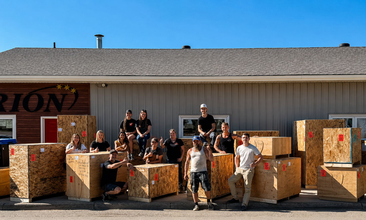Group of people posing among large cardboard crates outside a building with a sign reading 'RION' under a clear blue sky.