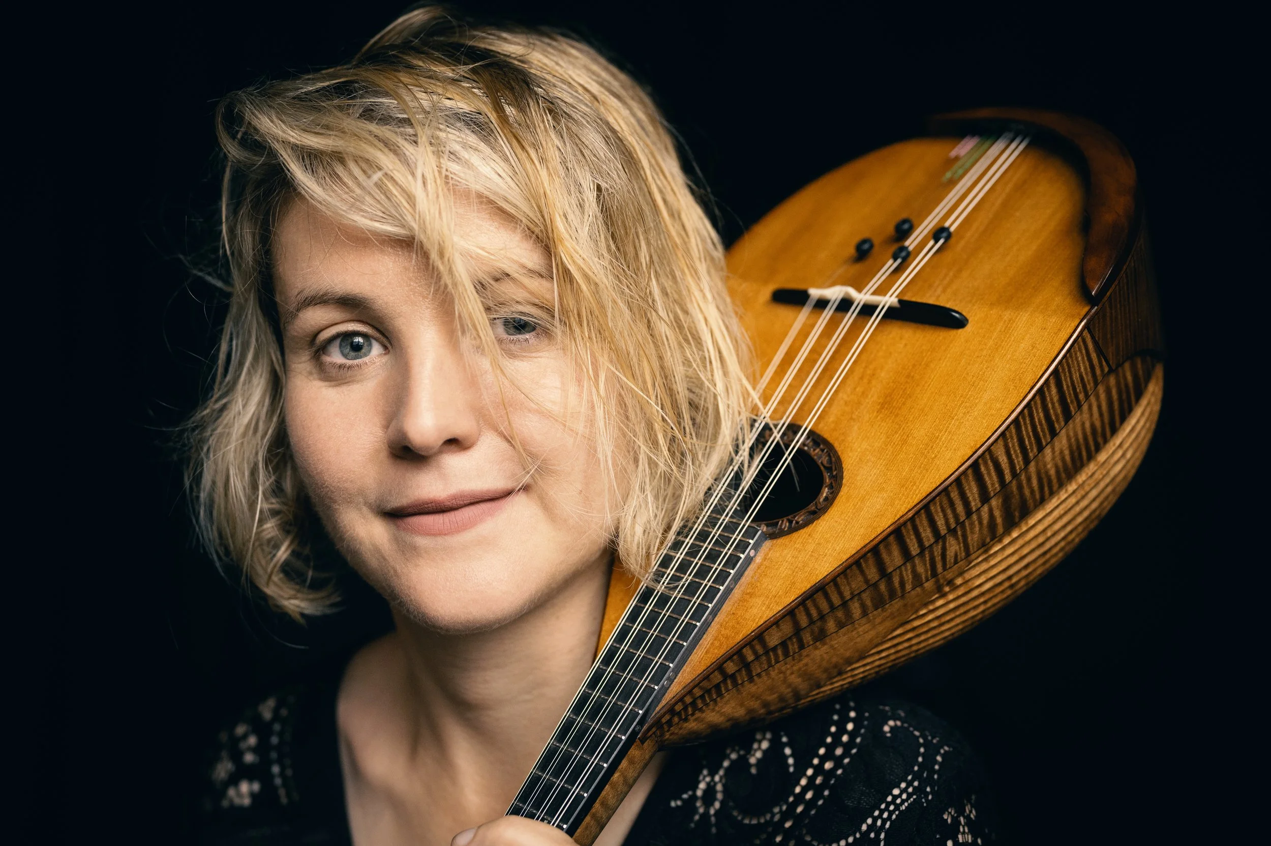 Close-up of a woman with blonde hair and blue eyes holding a wooden violin, against a black background.