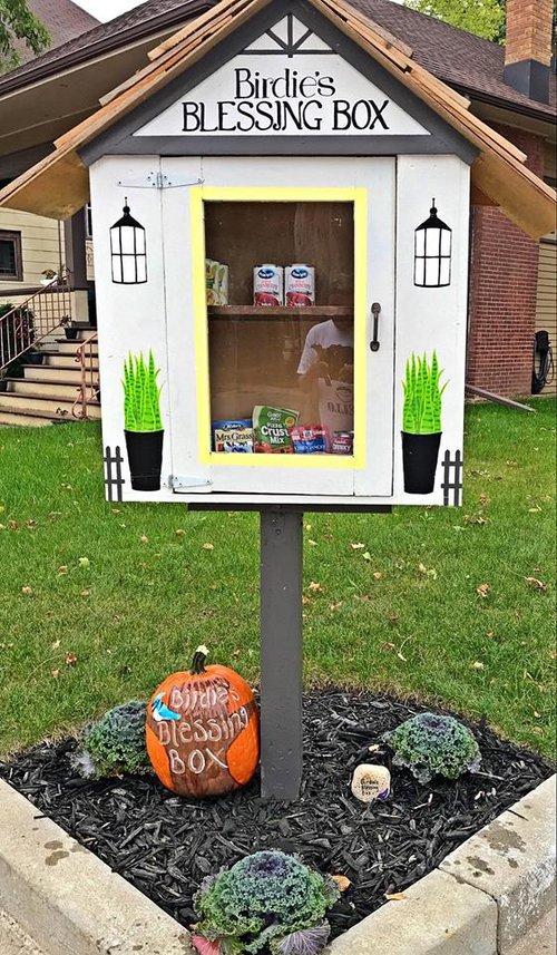 An outdoor wood cabinet with the appearance of a house. There's food inside which can be seen through the clear door. Text at the top of the cabinet reads "Birdie's Blessing Box"
