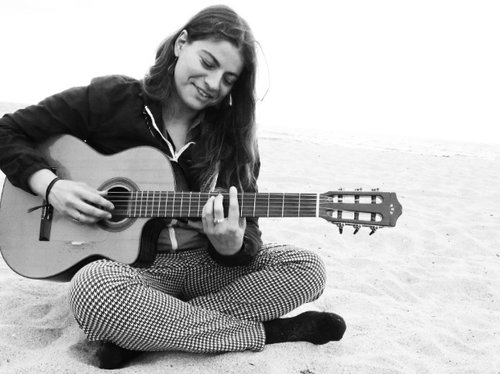 Lucy Morning star sitting crosslegged and playing guitar on a beach. The image is in black and white.