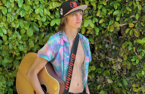BuhDihKuh in front of a hedge, wearing a blue shirt that's open, and a guitar across his chest. 