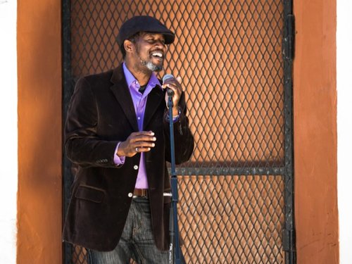 Chad in front of an orange wall and metal gate. He's wearing a bright violet shirt, and black velvet jacket and hat. He's smiling and singing into a microphone.