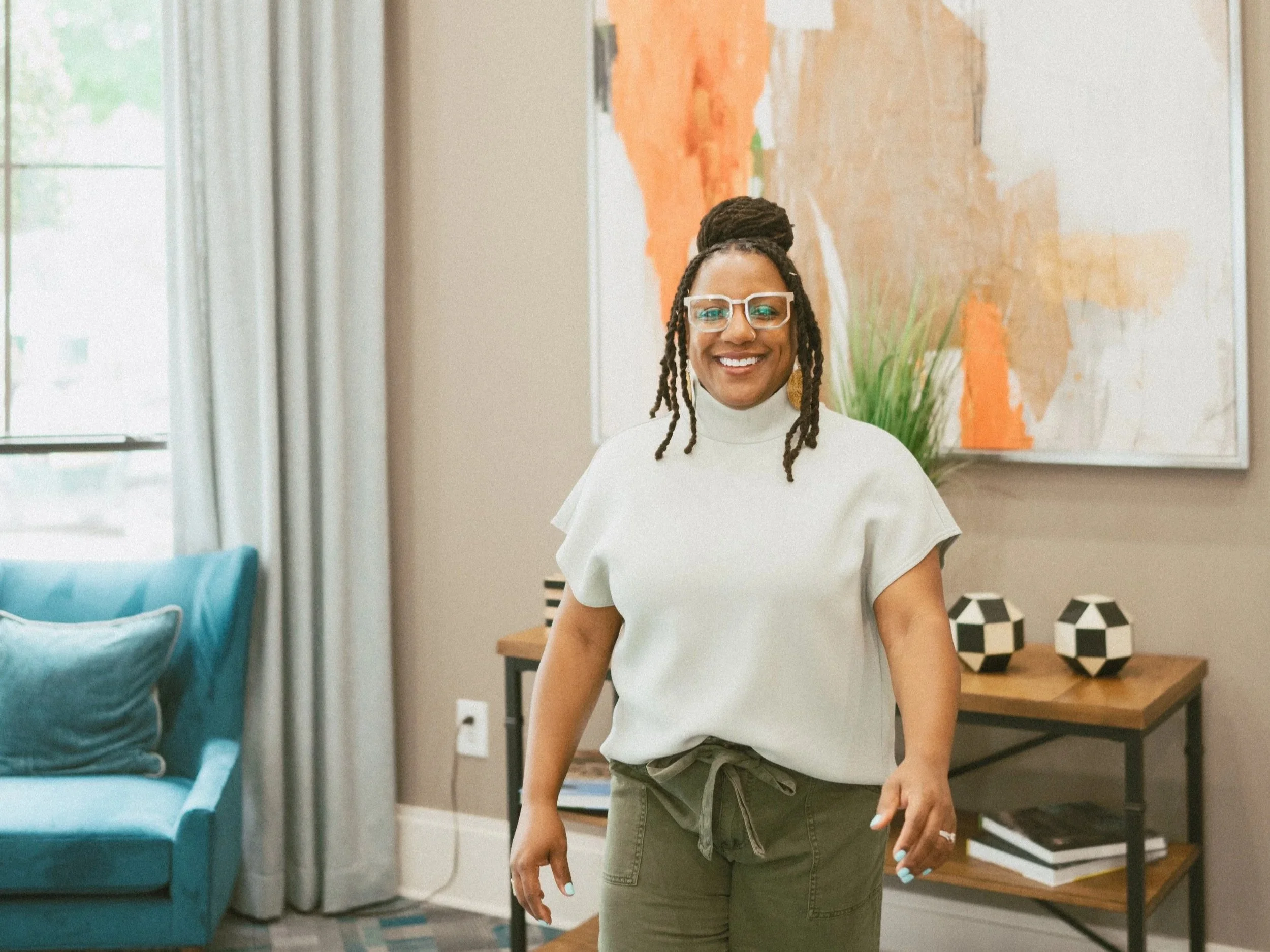 A smiling woman with dreadlocks and glasses standing in a modern living room with a teal sofa, large window, and abstract wall art.