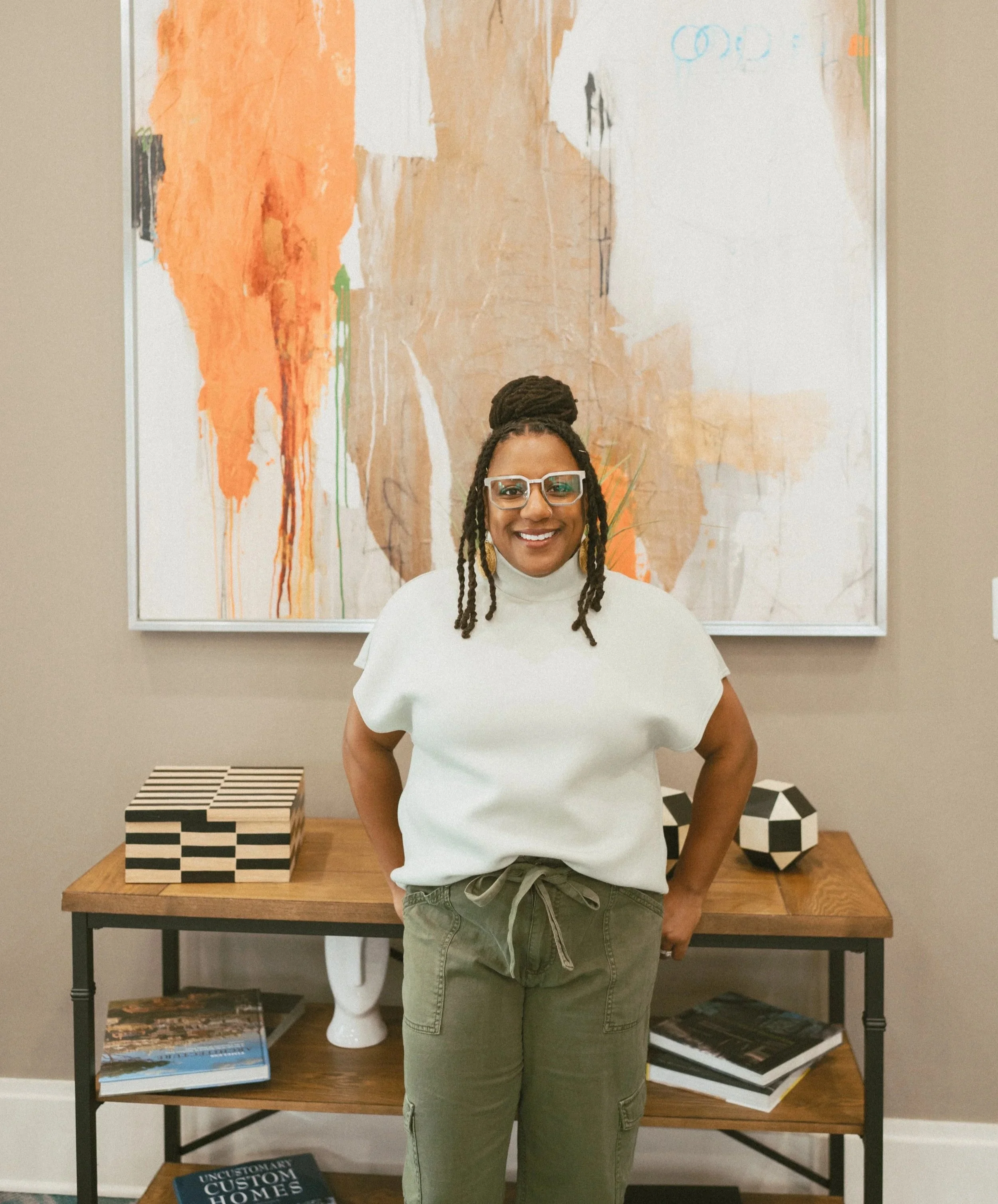 A smiling woman with glasses and dreadlocks standing in front of an abstract art piece in a gallery. She is wearing a white top and green pants.
