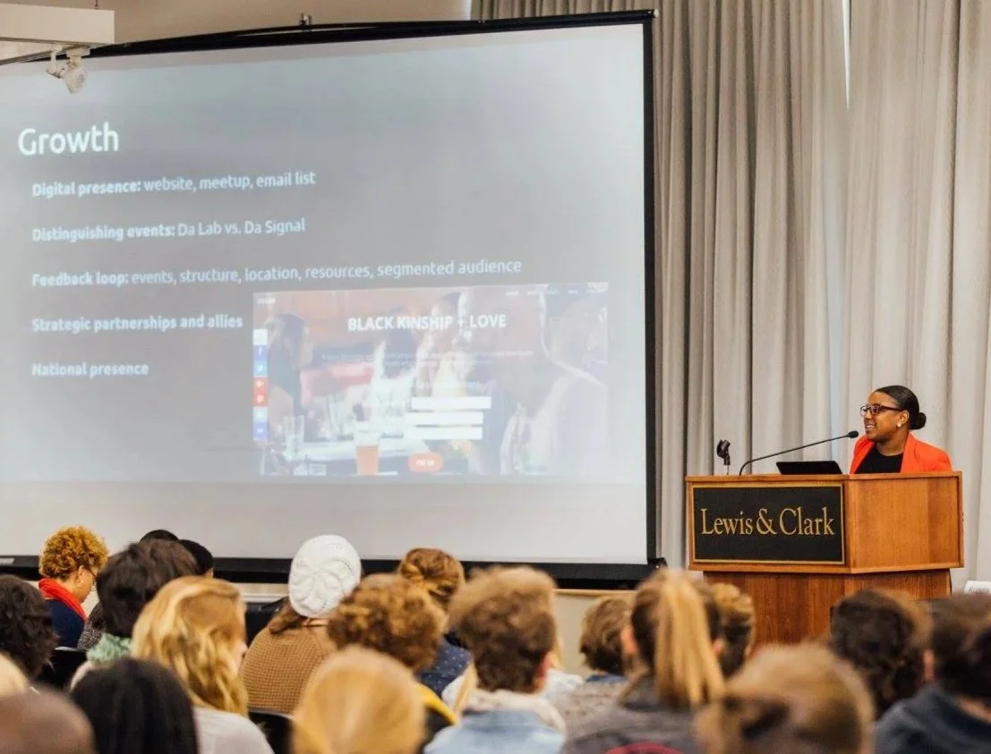 A woman in a red blazer is speaking at a podium with a sign that says "Lewis & Clark" to a large audience. A presentation slide titled "Growth" is projected on a screen behind her, listing points related to digital presence, events, feedback, partnerships, and national presence.
