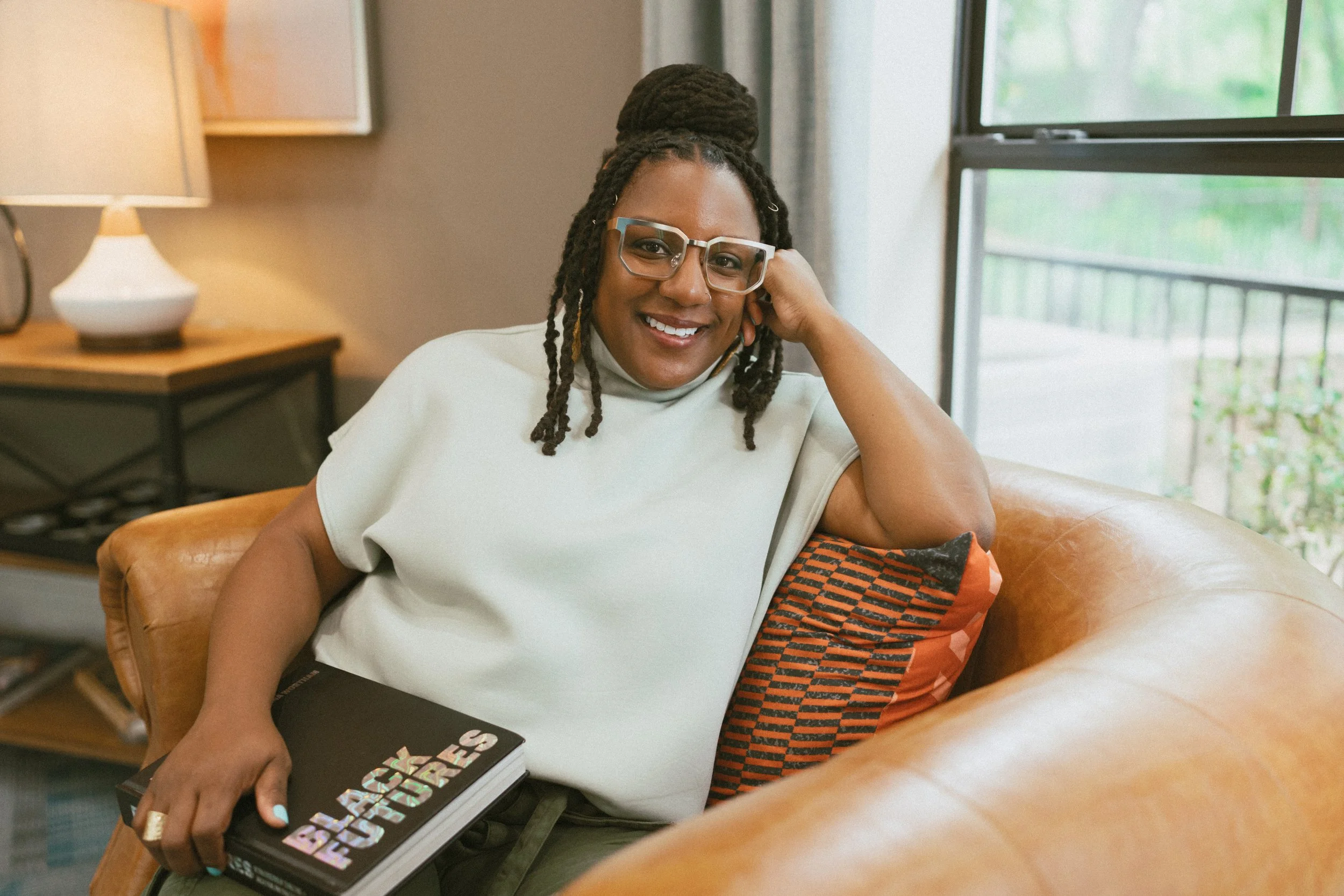 A woman with glasses and braided hair sitting on a leather couch in a living room, smiling, with a book in her hand, near a window with greenery outside.
