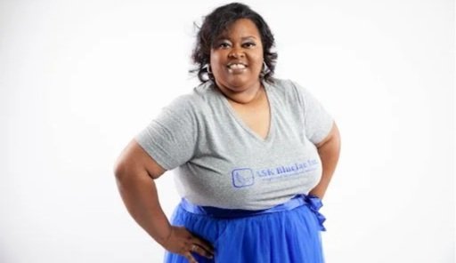 A woman smiling, wearing a gray t-shirt and blue skirt, standing against a plain white background.