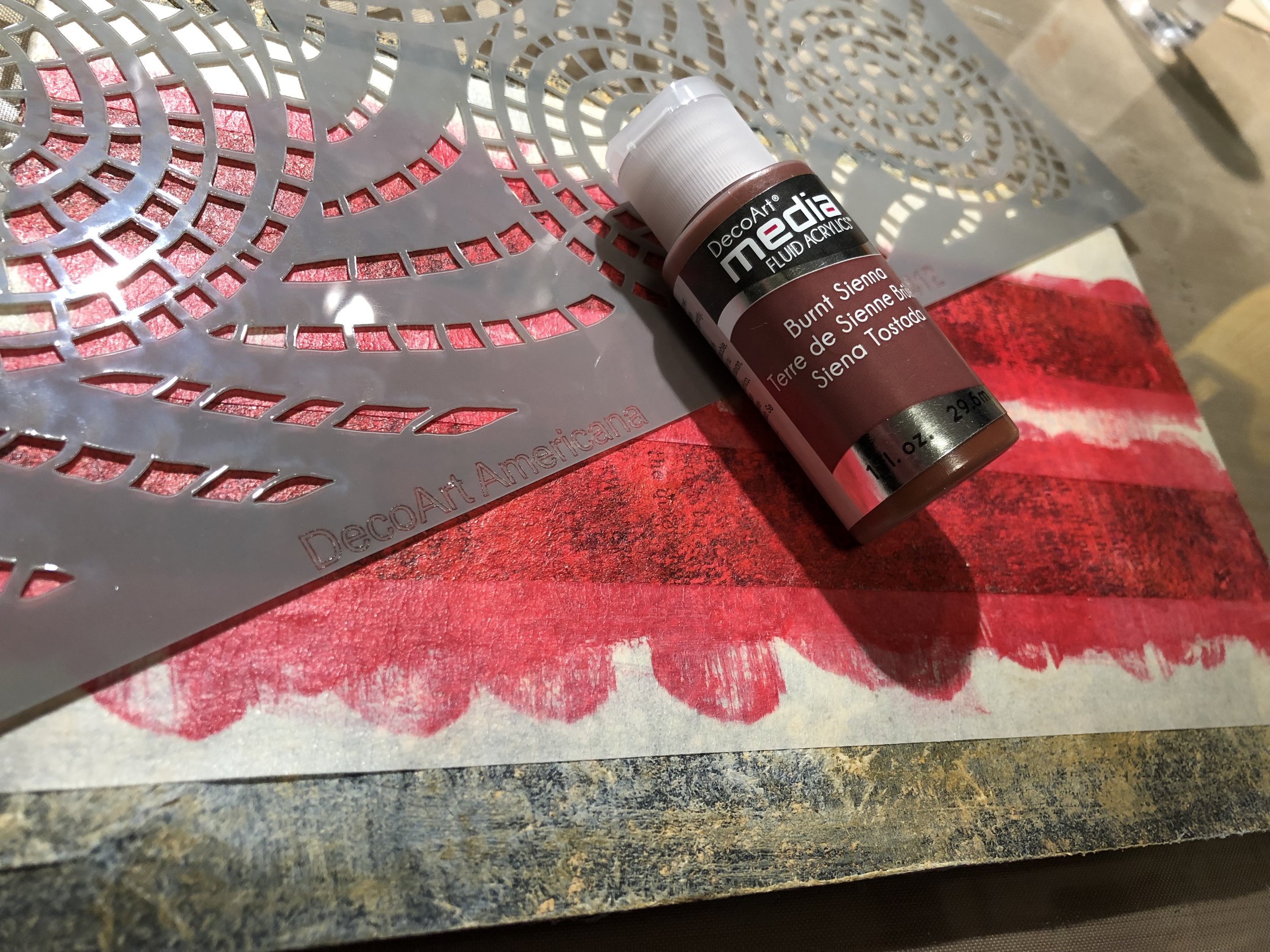 With a sponge and using DecoArt Americana’s ‘Segmented Swirls’ stencil, apply DecoArt Media Fluid Acrylic in ‘Burnt Sienna’ over the red stripes.