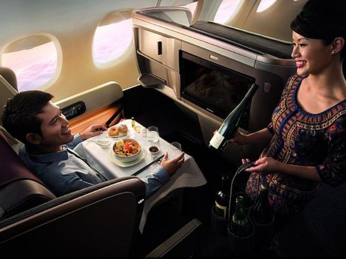 Onboard a Singapore Airlines plane a young man sits in a window seat, haute cuisine spread in front of him as a female flight attendant brings him wine with a smile.