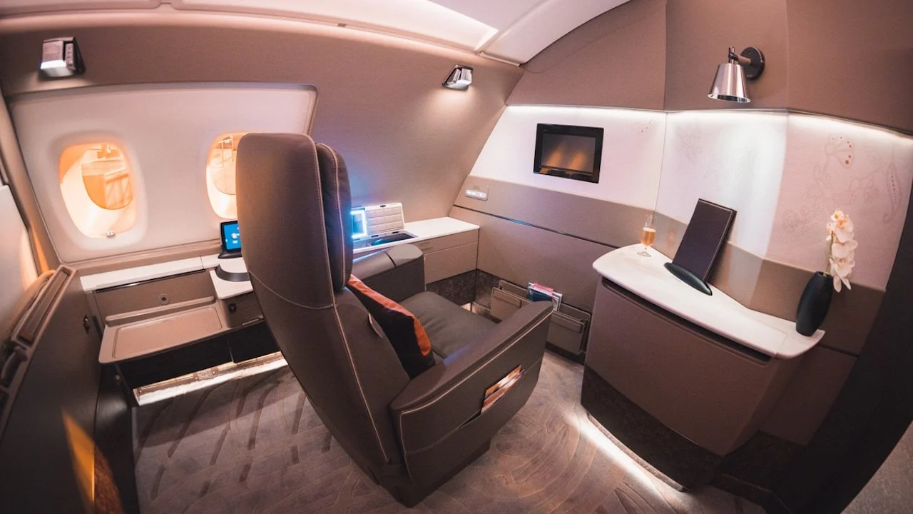 Interior of a luxury cabin onboard a Singapore Airlines plane. A comfy dark leather armchair, wall TV, elegant minimalist walls and carpet space. Champagne on a table top.