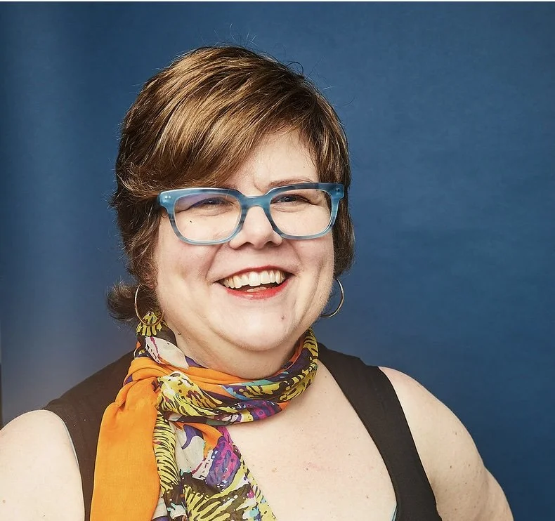 Jody Hankinson wearing a black top, orange patterned scarf, and bright blue glasses