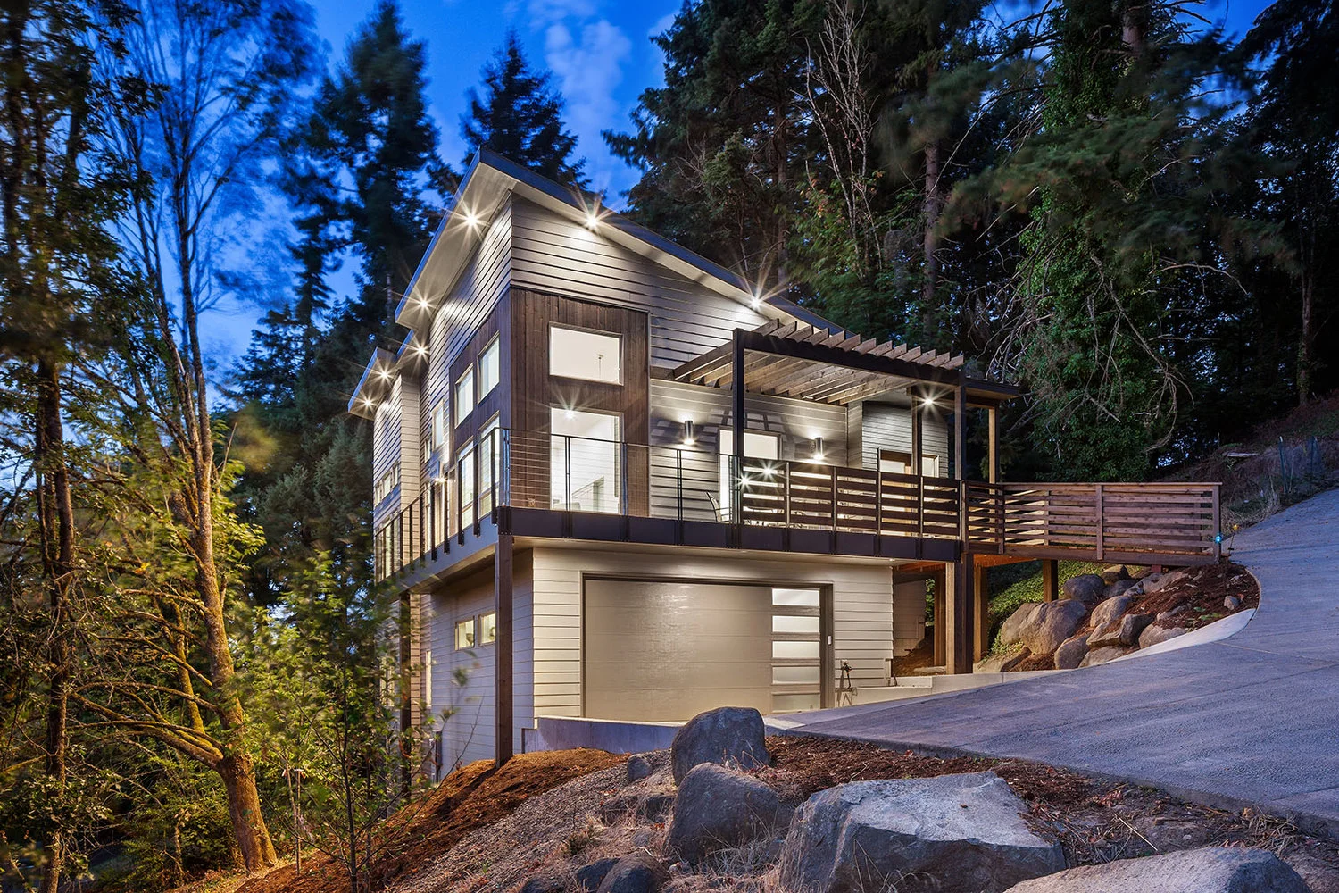 A modern hillside home lit up at dusk among tall firs