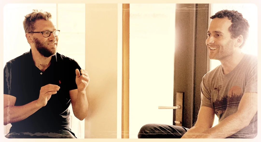 Two men sitting and talking, one wearing glasses and a black shirt, the other in a graphic tee. They are smiling and engaged in conversation, with a bright window in the background.