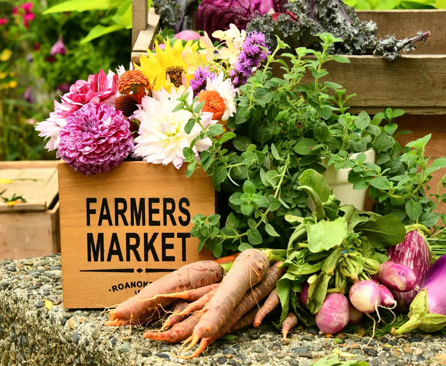 A wooden sign reading 'Farmers Market' surrounded by fresh vegetables including carrots and radishes, and colorful flowers at an outdoor market.