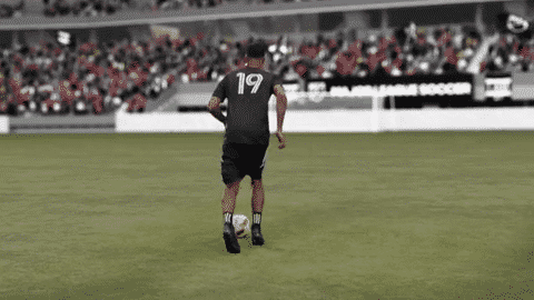A soccer player wearing a black uniform with the number 19 on the back, running on a soccer field with a crowd in the stands.