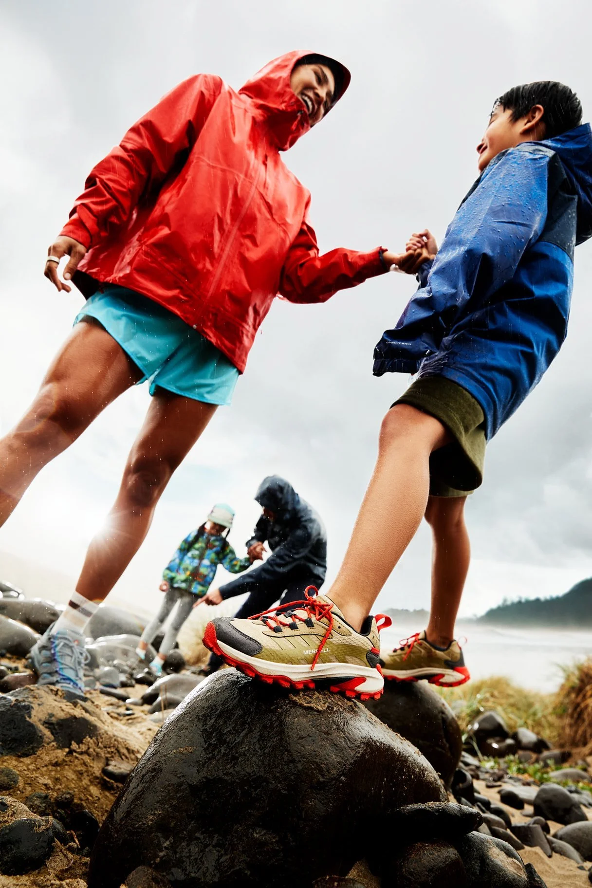 People outdoors in rainy weather, two children holding hands and balancing on rocks, wearing rain jackets and hiking shoes, with others in the background near water.