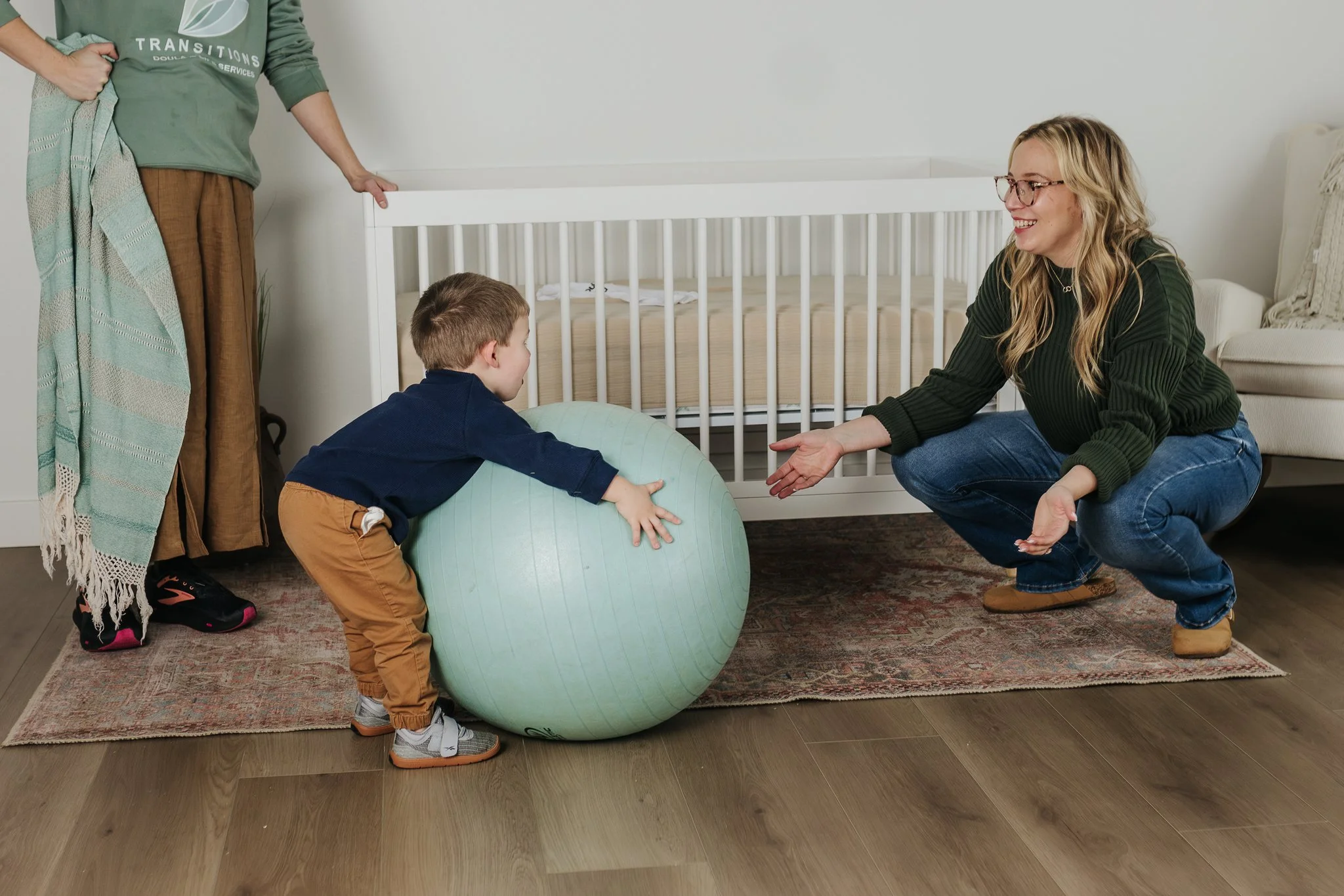A woman plays with a toddler and a birth ball in front of a crib.