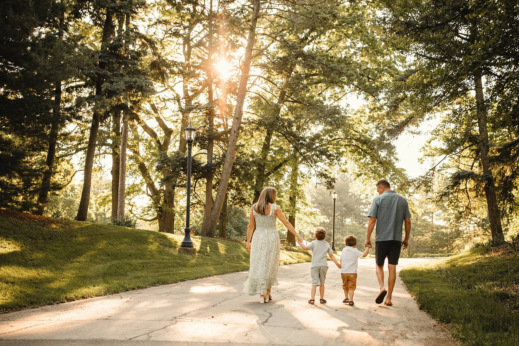 Family of four walking hand in hand on a sunny park path during sunset, surrounded by tall trees and green grass during an outdoor family photoshoot in Oak Brook, IL.