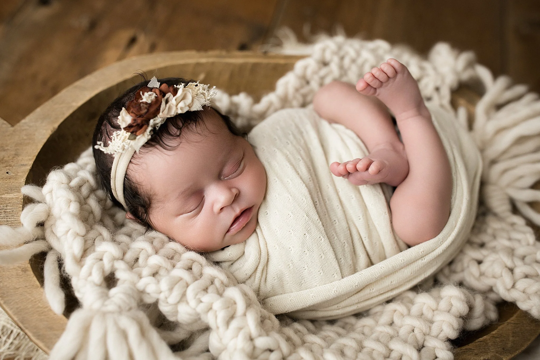 A sleeping baby with dark hair and light skin, wearing a cream-colored outfit and a floral headband, resting on a chunky knit blanket inside a wooden bowl with newborn photography Naperville.