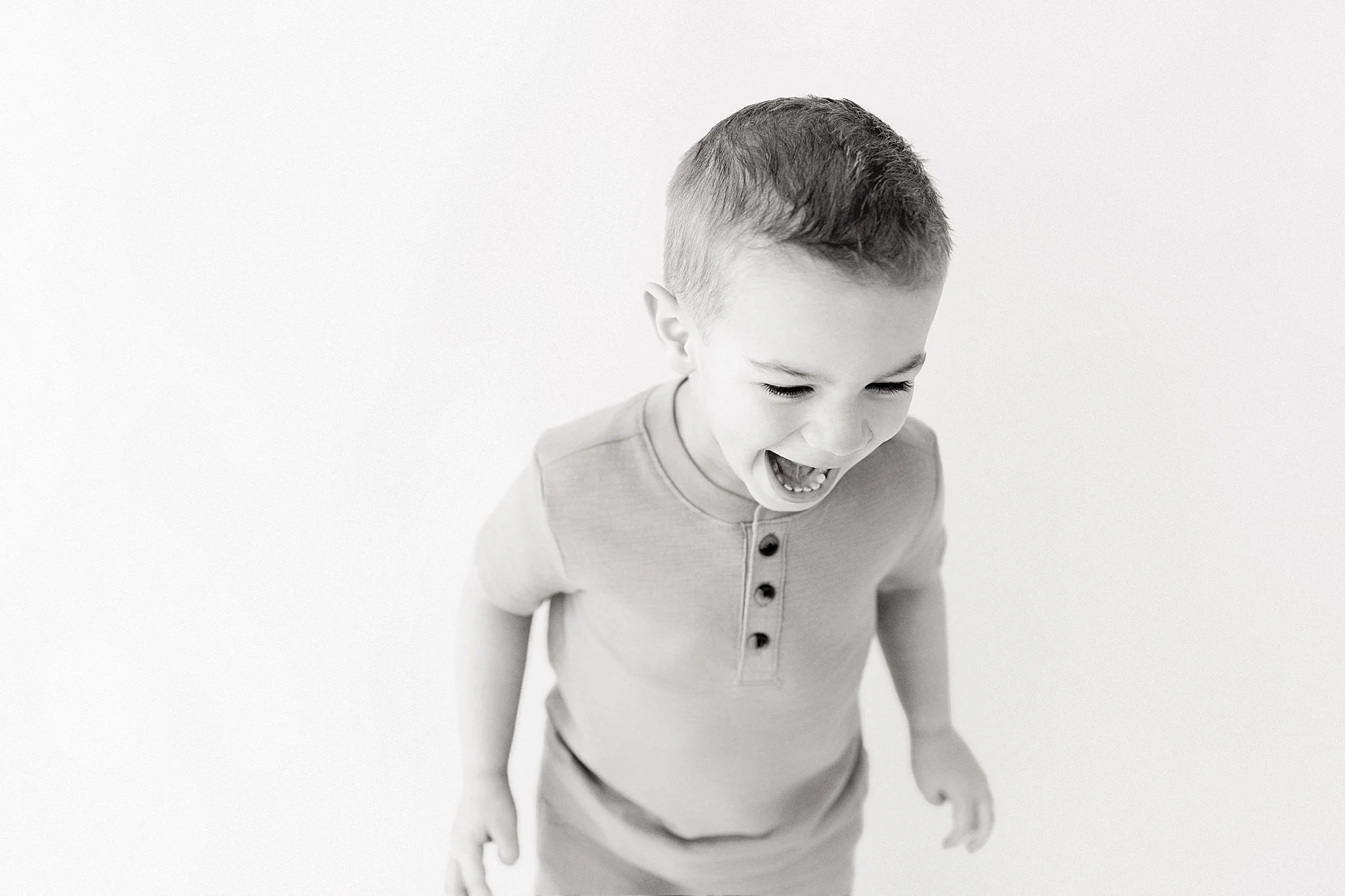 A young boy with short hair laughing with his mouth open, wearing a light-colored shirt with buttons, looking downward, against a plain light background with Marie Lopez Naperville photographer.