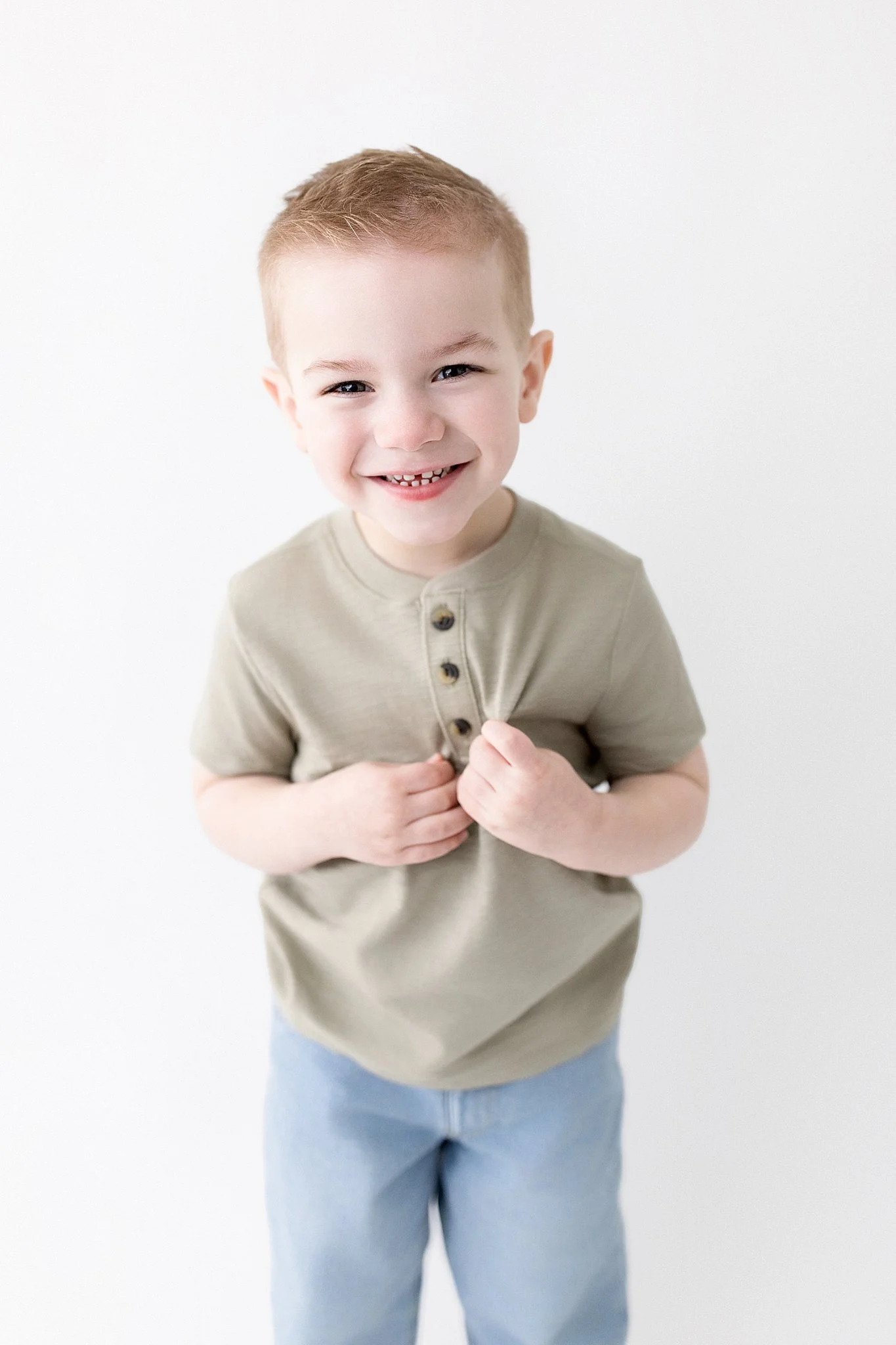 Young boy with short red hair smiling, wearing a beige shirt and light blue pants, standing against a white background during a kids studio photography session in Naperville.