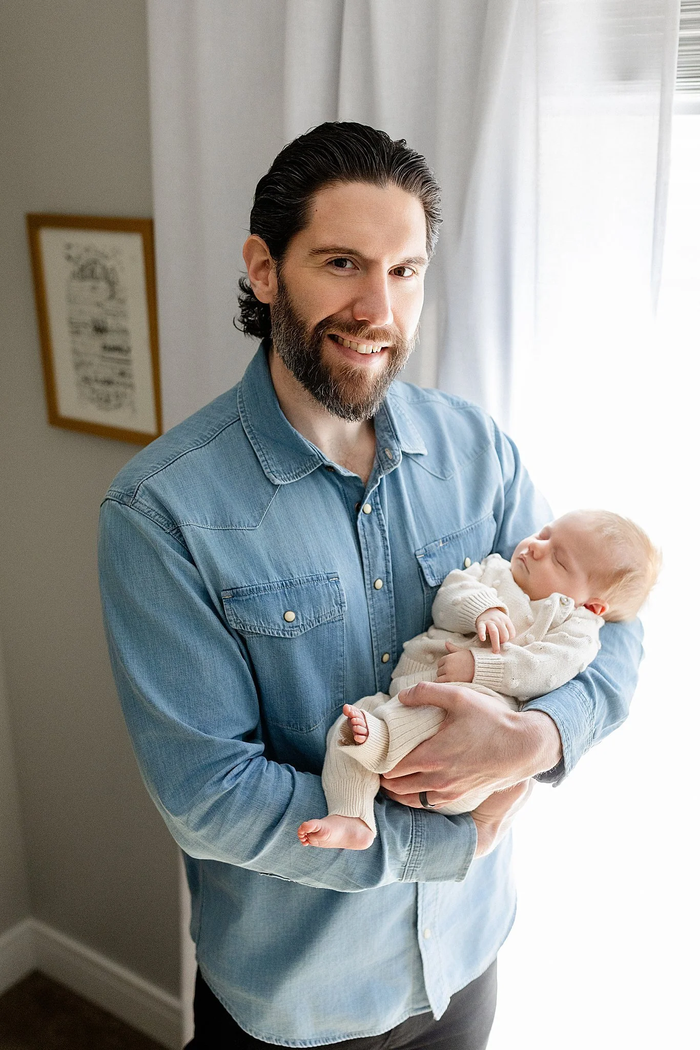 A man with dark hair and a beard holding a sleeping baby, standing in front of a window with white curtains and a framed picture on the wall with Naperville photographers.