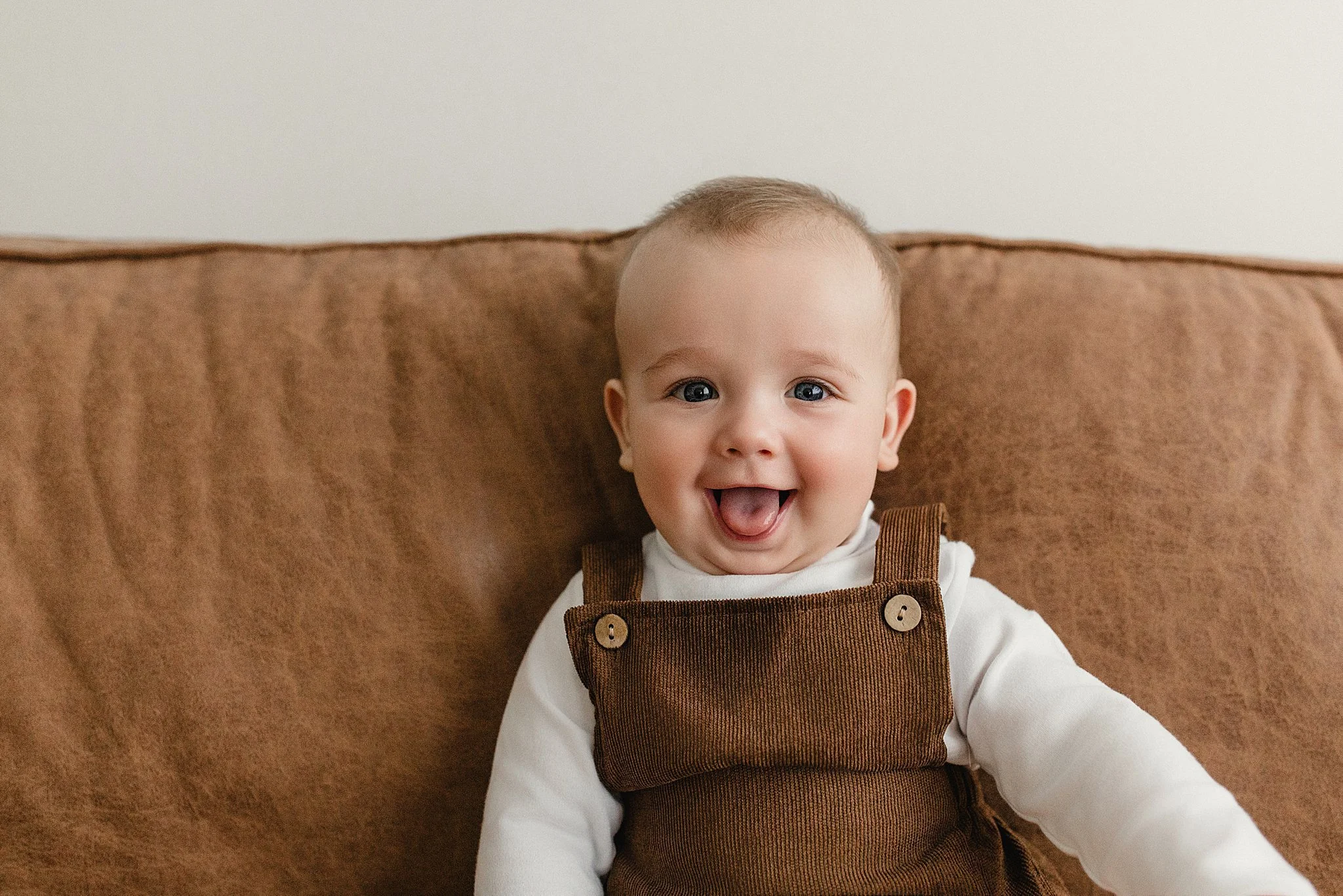 Cute baby with bright blue eyes and a big smile, wearing brown overalls and a white long-sleeve shirt, sitting on a brown couch with a plain wall background with Ally and B Photography in downtown Naperville, IL.