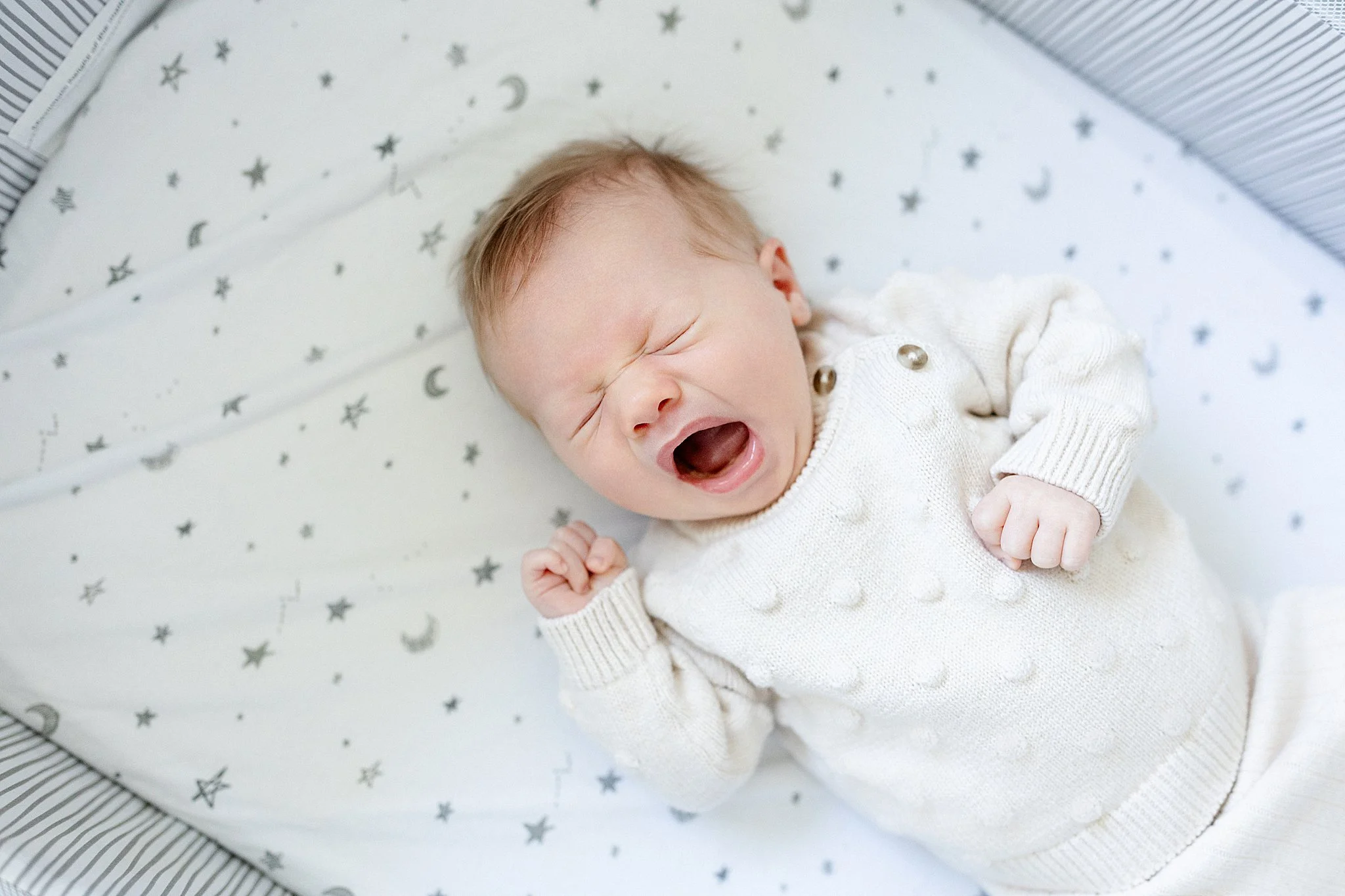 A baby with light skin and brown hair yawning, lying on a star and moon patterned white bed sheet inside a bed with a black and white striped edge.