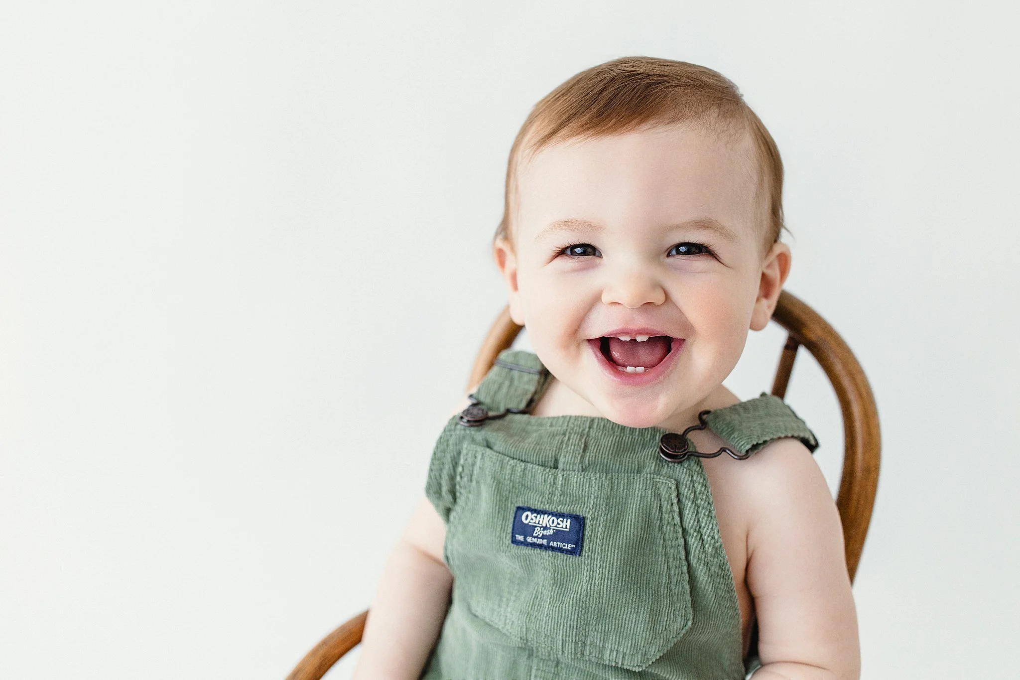 Smiling baby with light brown hair and blue eyes, wearing green OshKosh overalls, sitting on a wooden chair, against a plain white background for baby photoshoot in Naperville, IL.