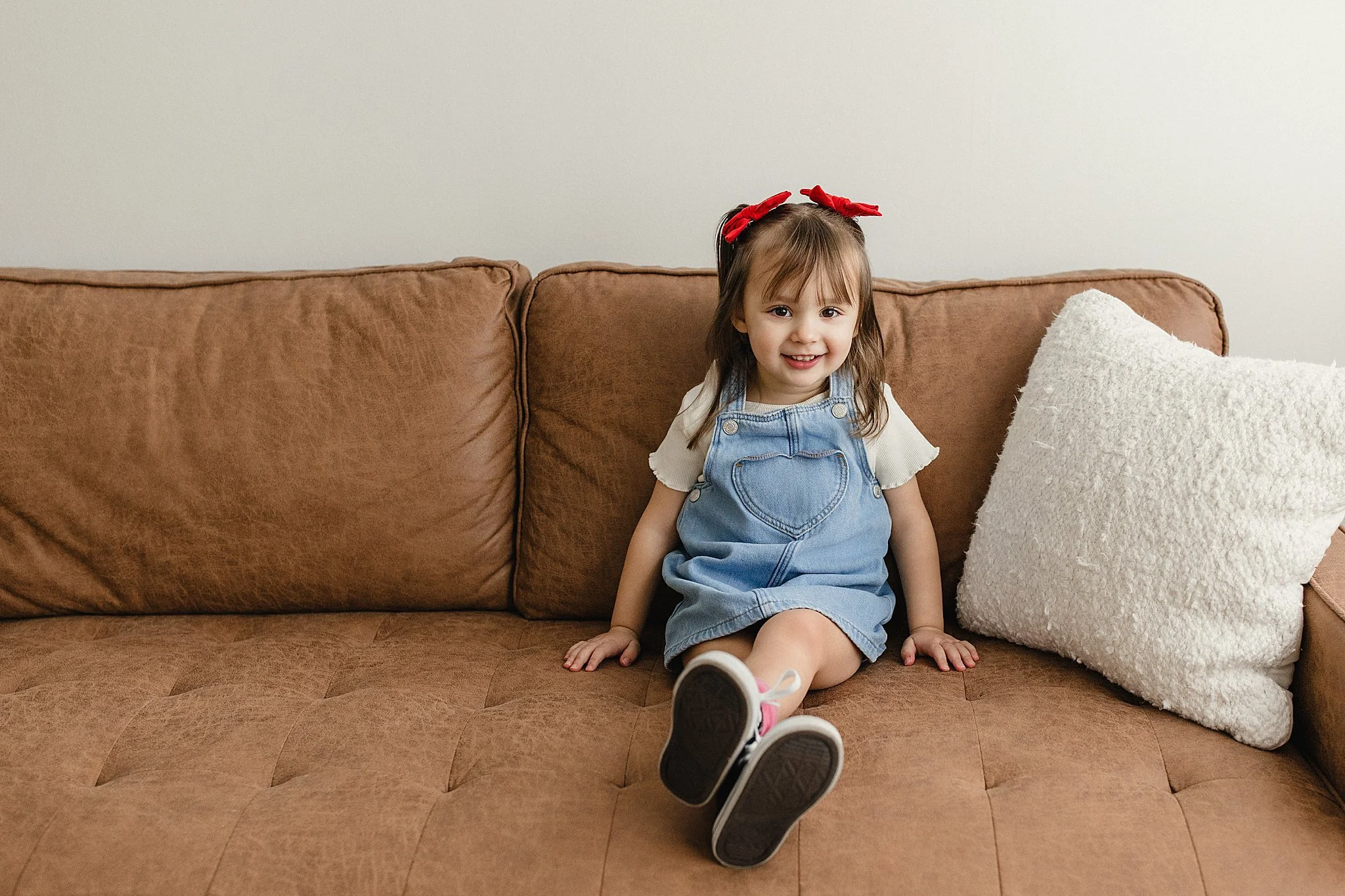 A smiling young girl with brown hair seated on a brown sofa, wearing a white t-shirt, denim overalls with a heart-shaped pocket, pink and white sneakers, and a red headband with a bow, next to a white textured pillow during kids photoshoot near me.