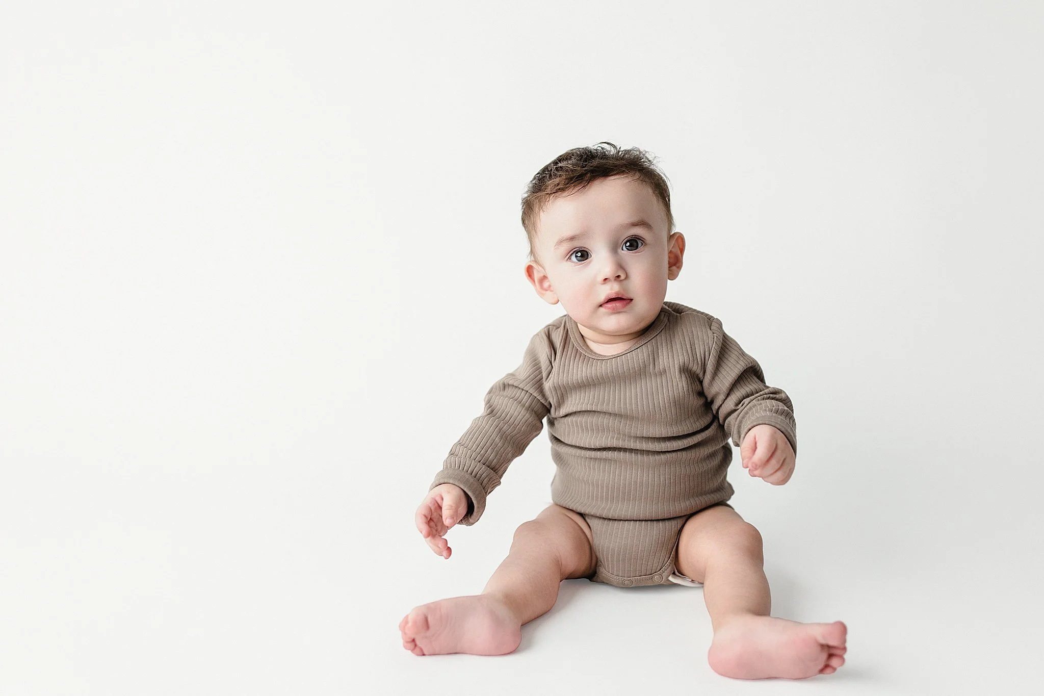 A baby sitting on the floor with a surprised expression, wearing a beige long-sleeve shirt and diaper, against a plain white background.