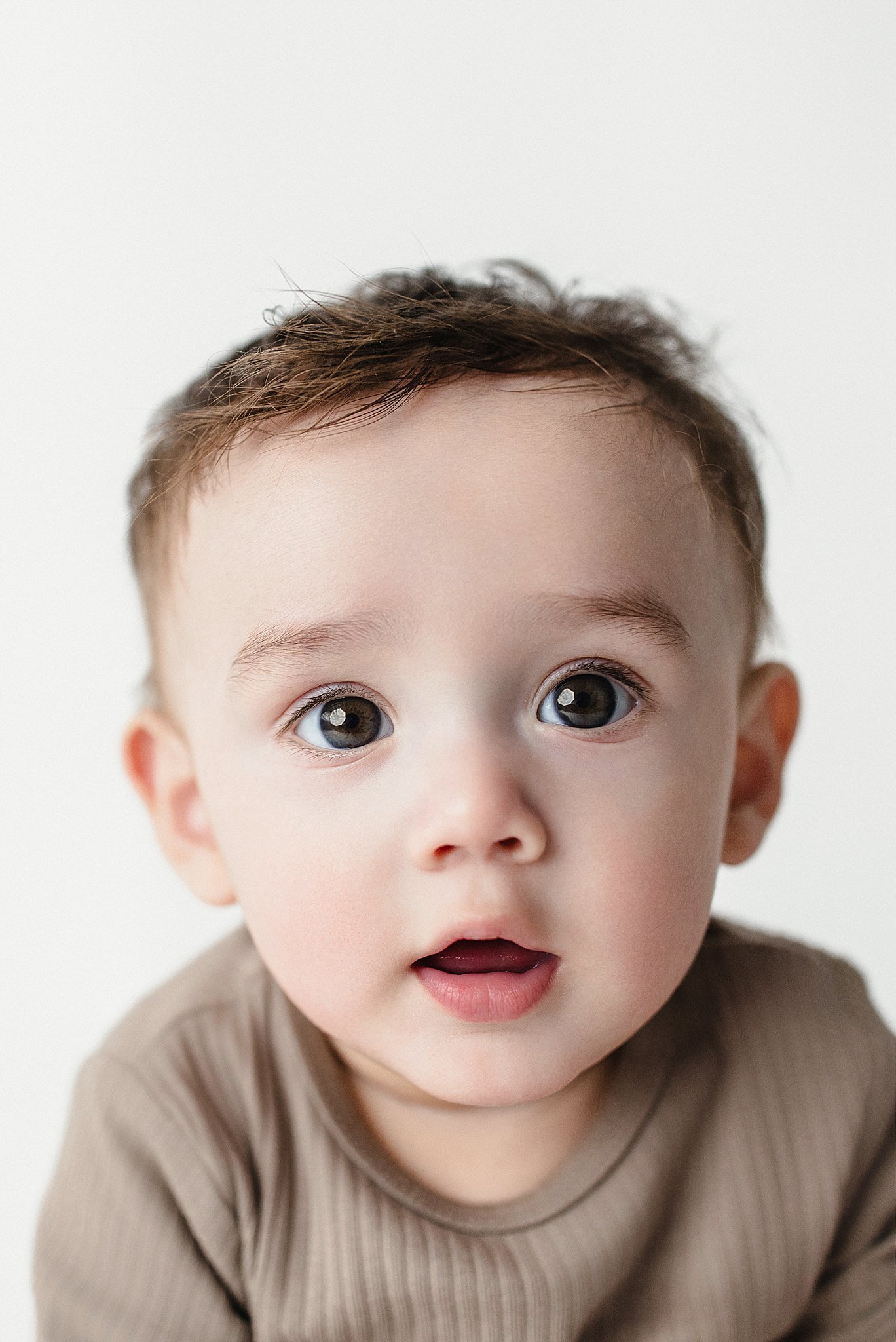 Close-up of a young child with big, expressive blue eyes and light brown hair, wearing a beige shirt, against a white background with best baby photographer in Naperville, IL.