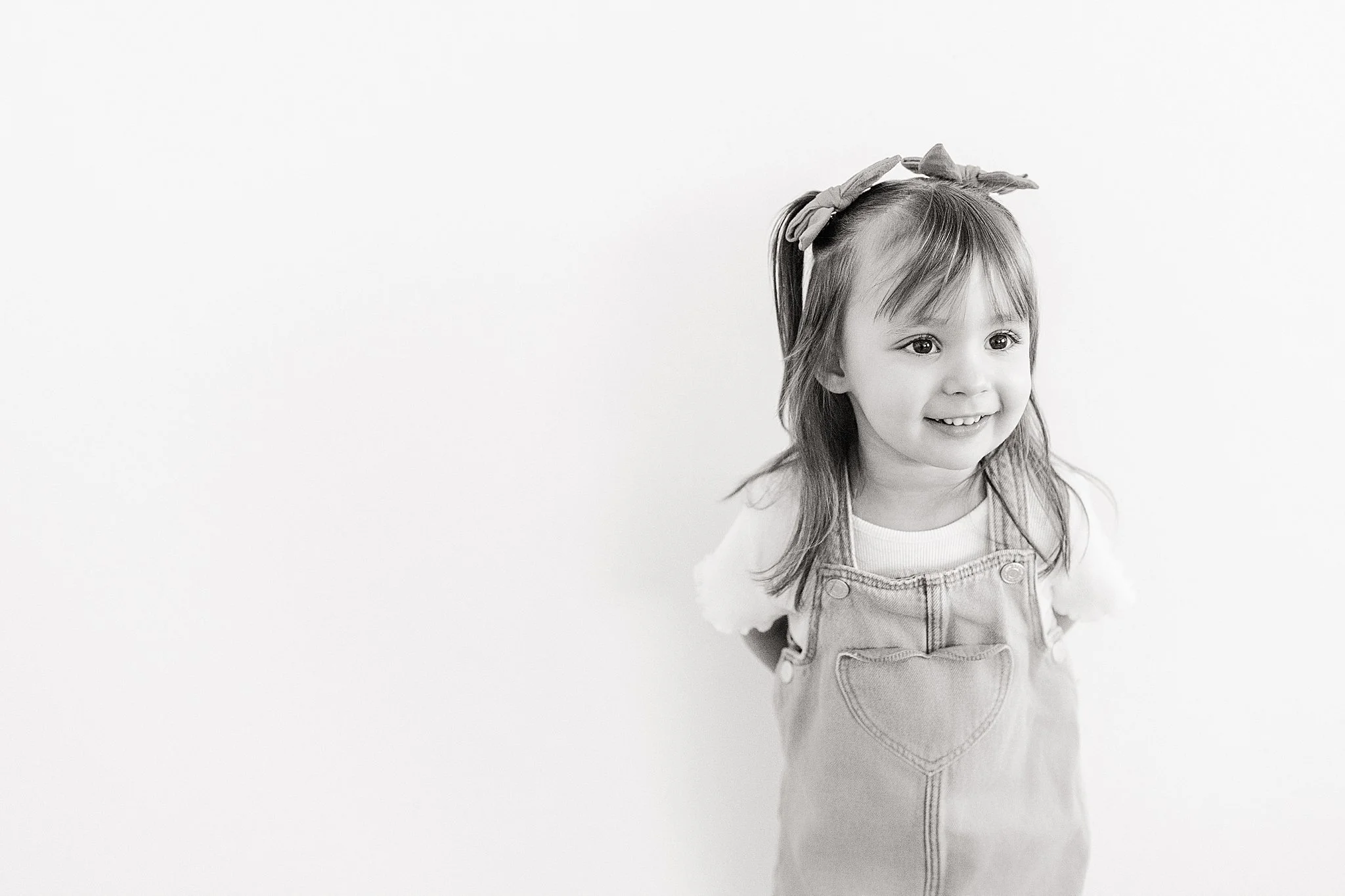 A young girl with a bow in her hair, smiling, wearing a denim overall and a t-shirt, standing against a plain white background for children's photography near me.