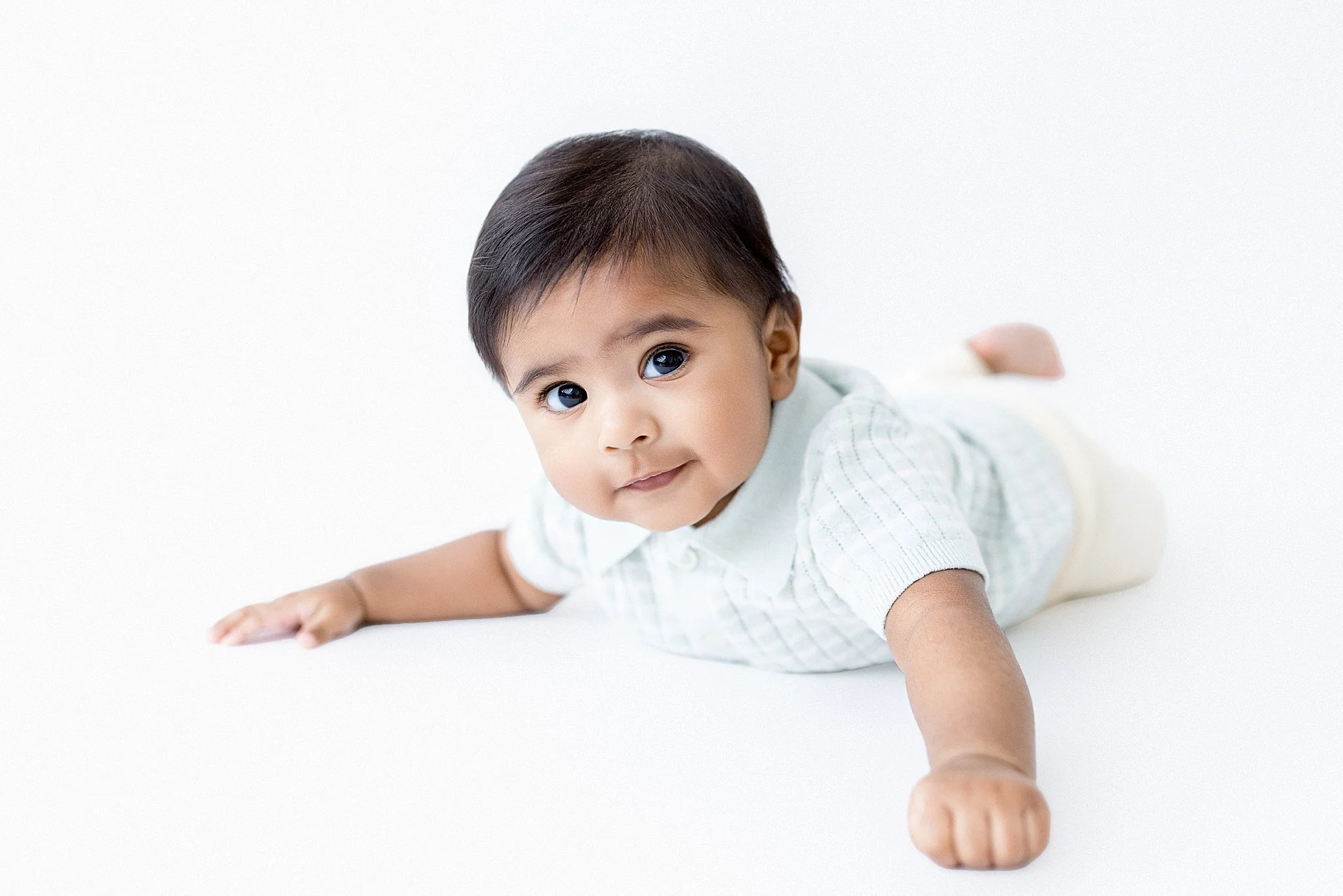 A baby lying on a white surface, looking at the camera with a gentle smile, wearing a white outfit with a collar during a baby photoshoot with Ally and B Photography.
