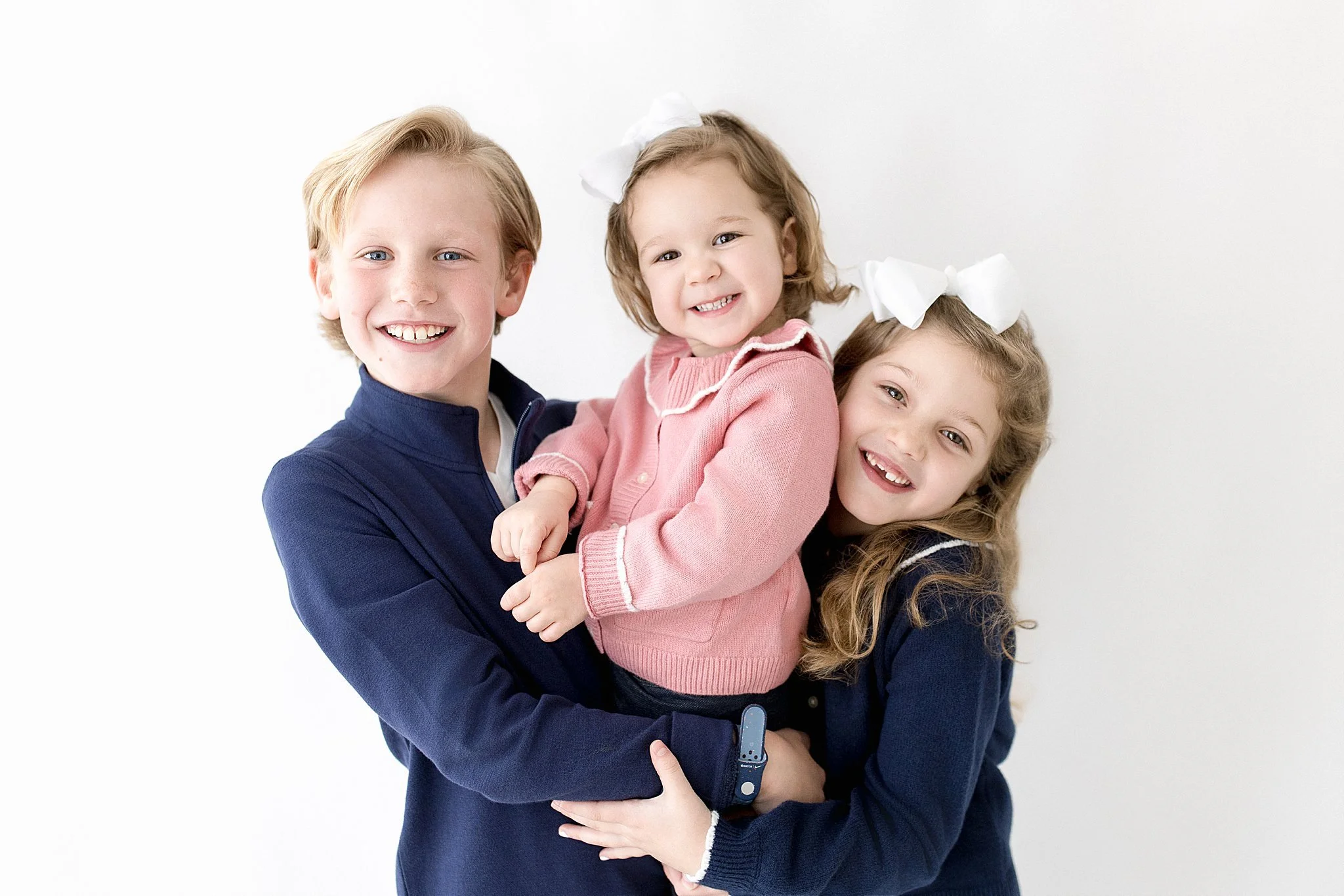 Three children, a boy and two girls, smiling and hugging each other against a plain white background with the best kids photographer in Naperville, IL.