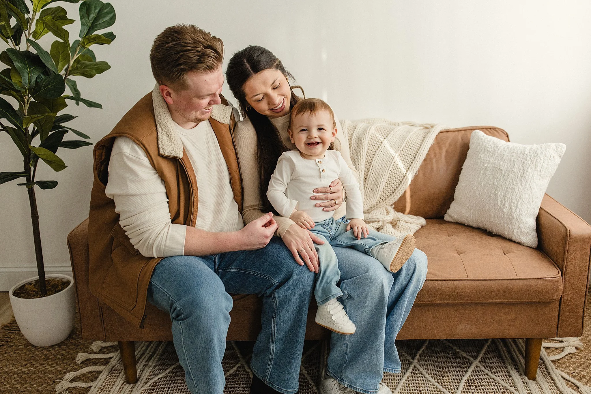 A happy family of three sitting together on a brown sofa in a cozy living room, with a potted plant beside them and a textured white cushion and blanket on the sofa during a studio lifestyle family photo session with Ally and B Photography.