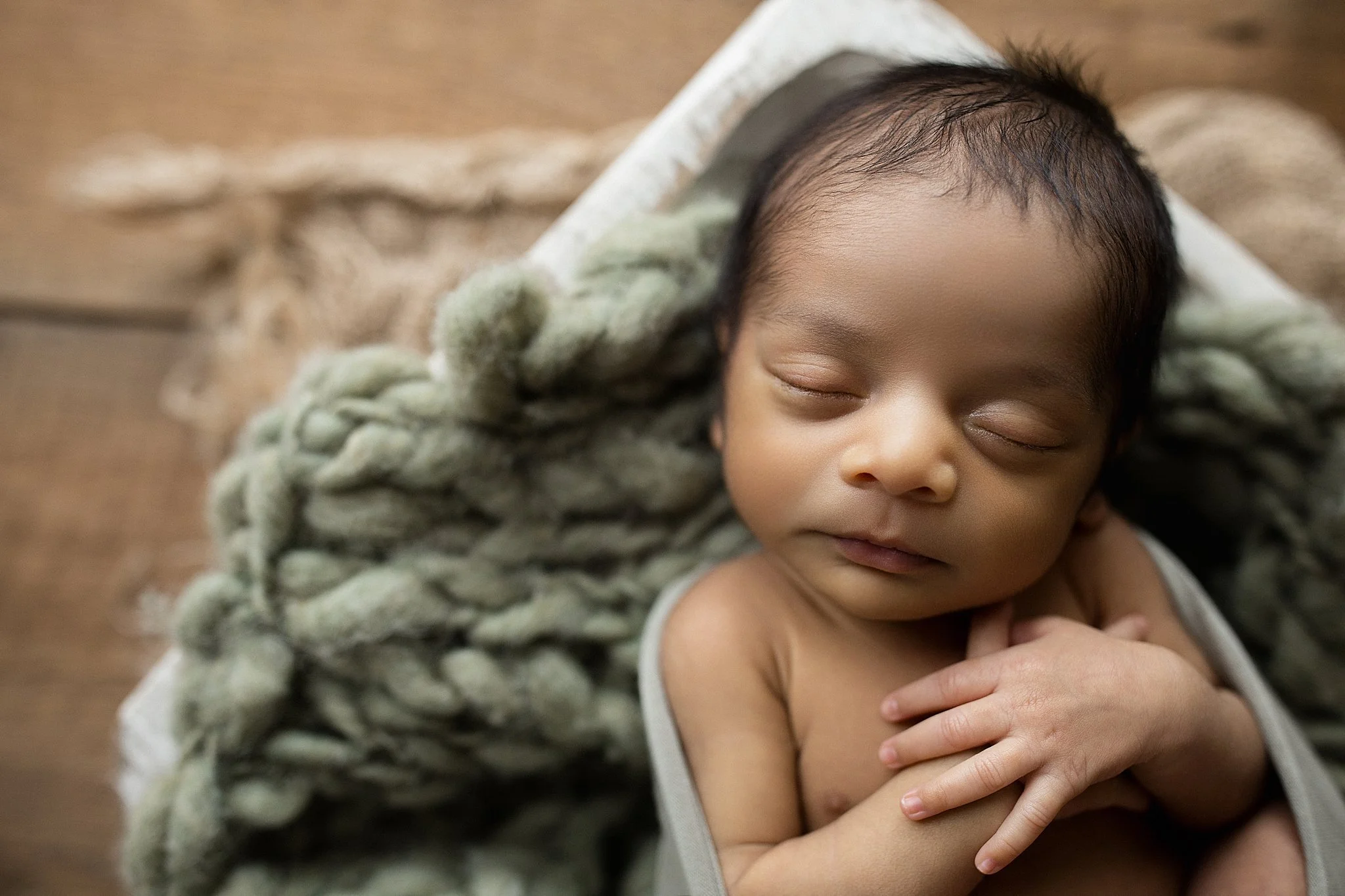 A sleeping newborn baby with dark hair, resting on a soft, chunky knit blanket on a wooden surface during posed newborn photography with Ally and B Photography in Naperville, IL.
