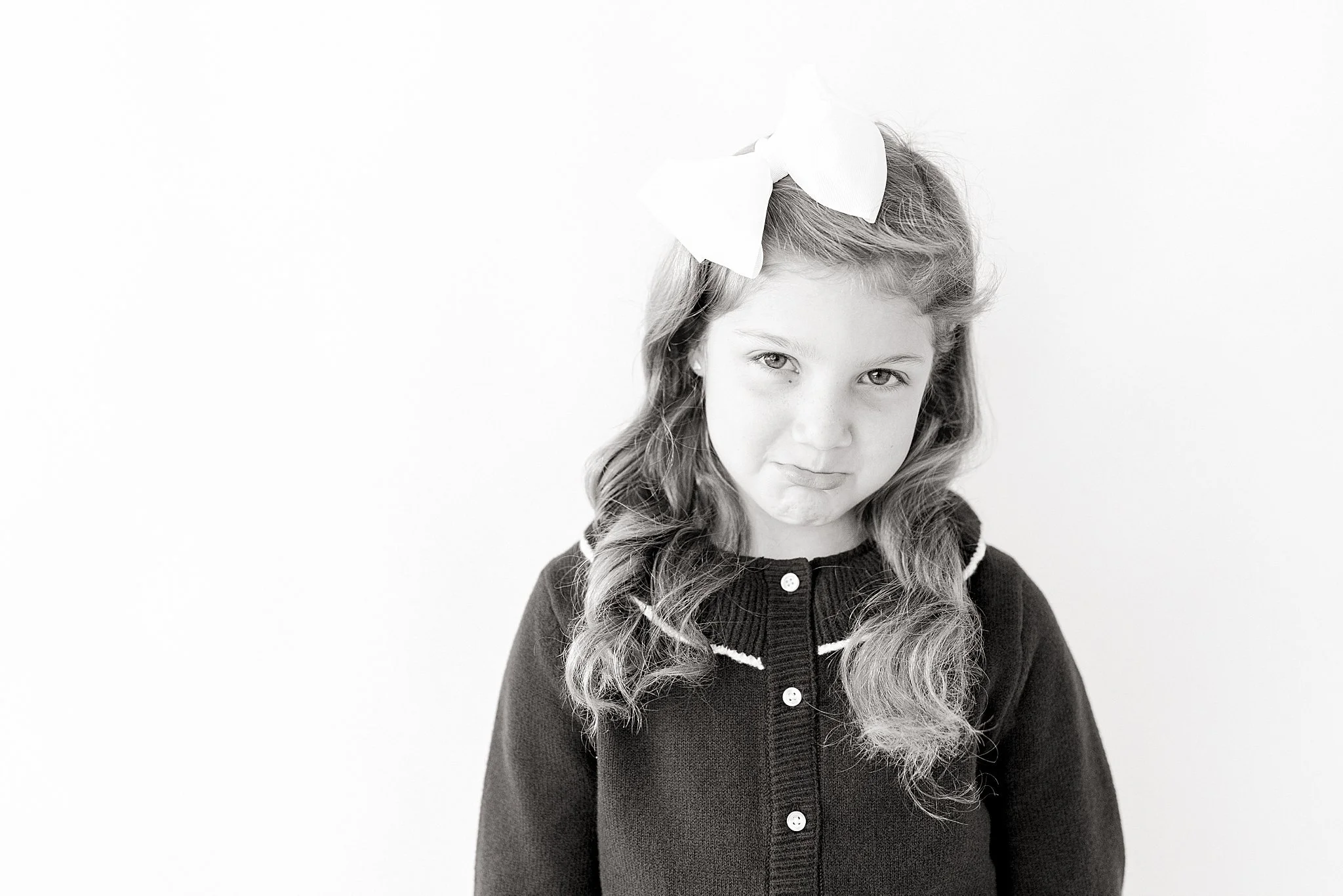 A young girl with long curly hair wearing a dark sweater and a large white bow in her hair, making a displeased facial expression against a plain light background with the best children's photographer in Naperville, IL.