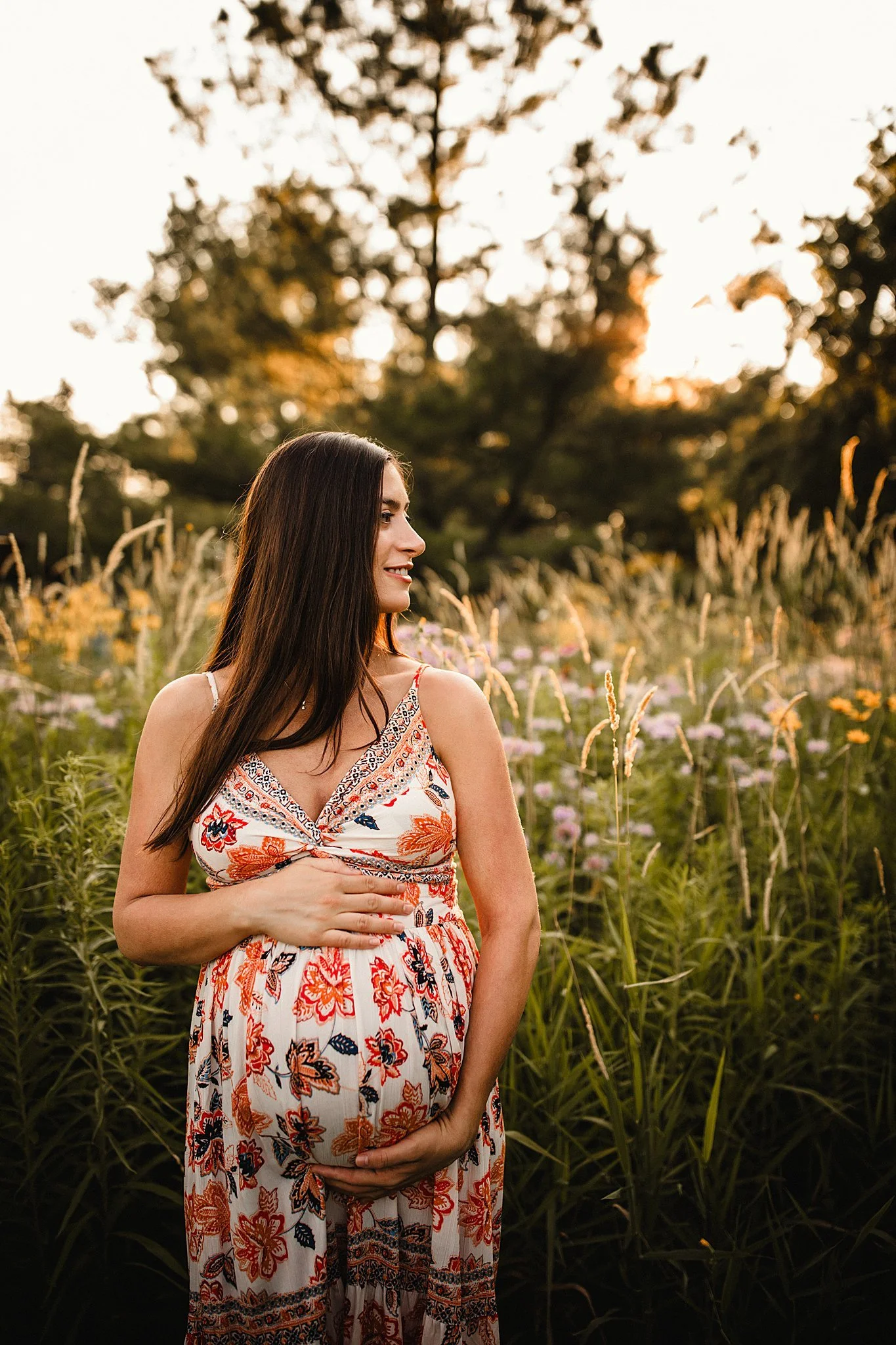A pregnant woman in a floral dress standing in a field of tall grass and wildflowers at sunset, holding her belly and smiling during a golden hour outdoor maternity session with Ally and B Photography.