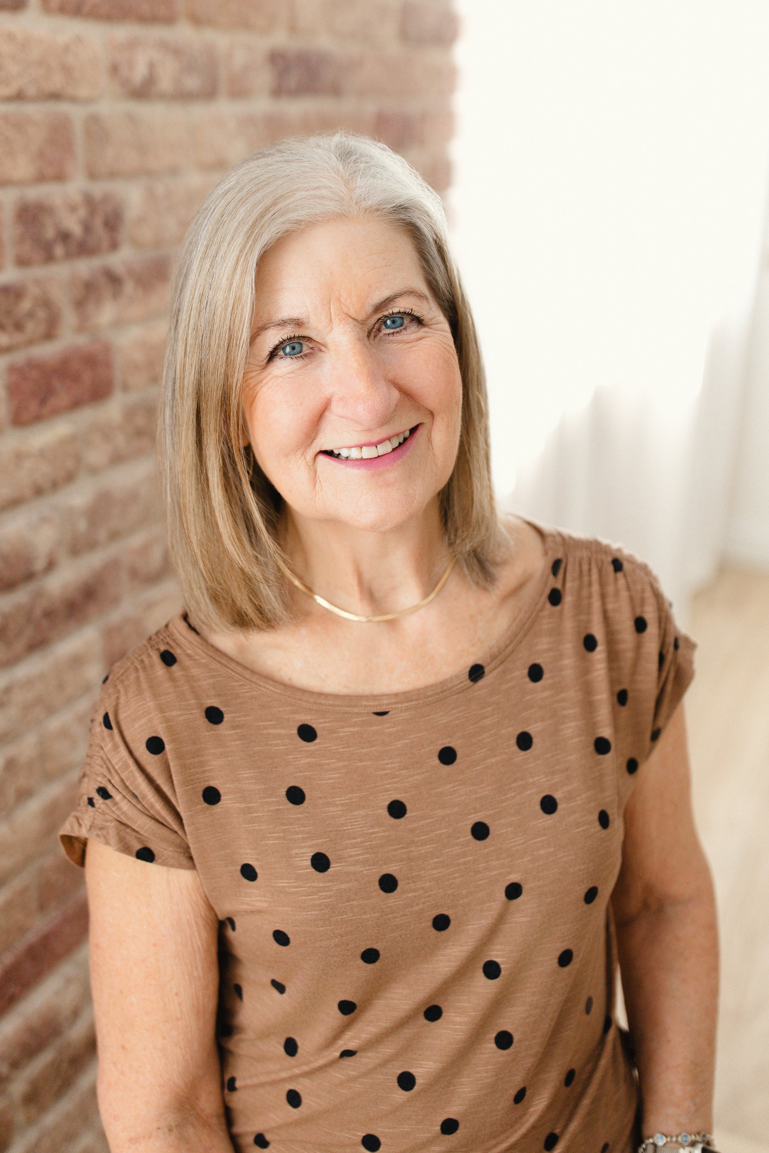 An elderly woman with shoulder-length gray hair and blue eyes, smiling, wearing a brown polka dot top and a gold necklace, standing near a brick wall in a well-lit room for headshots with Ally and B Photography.