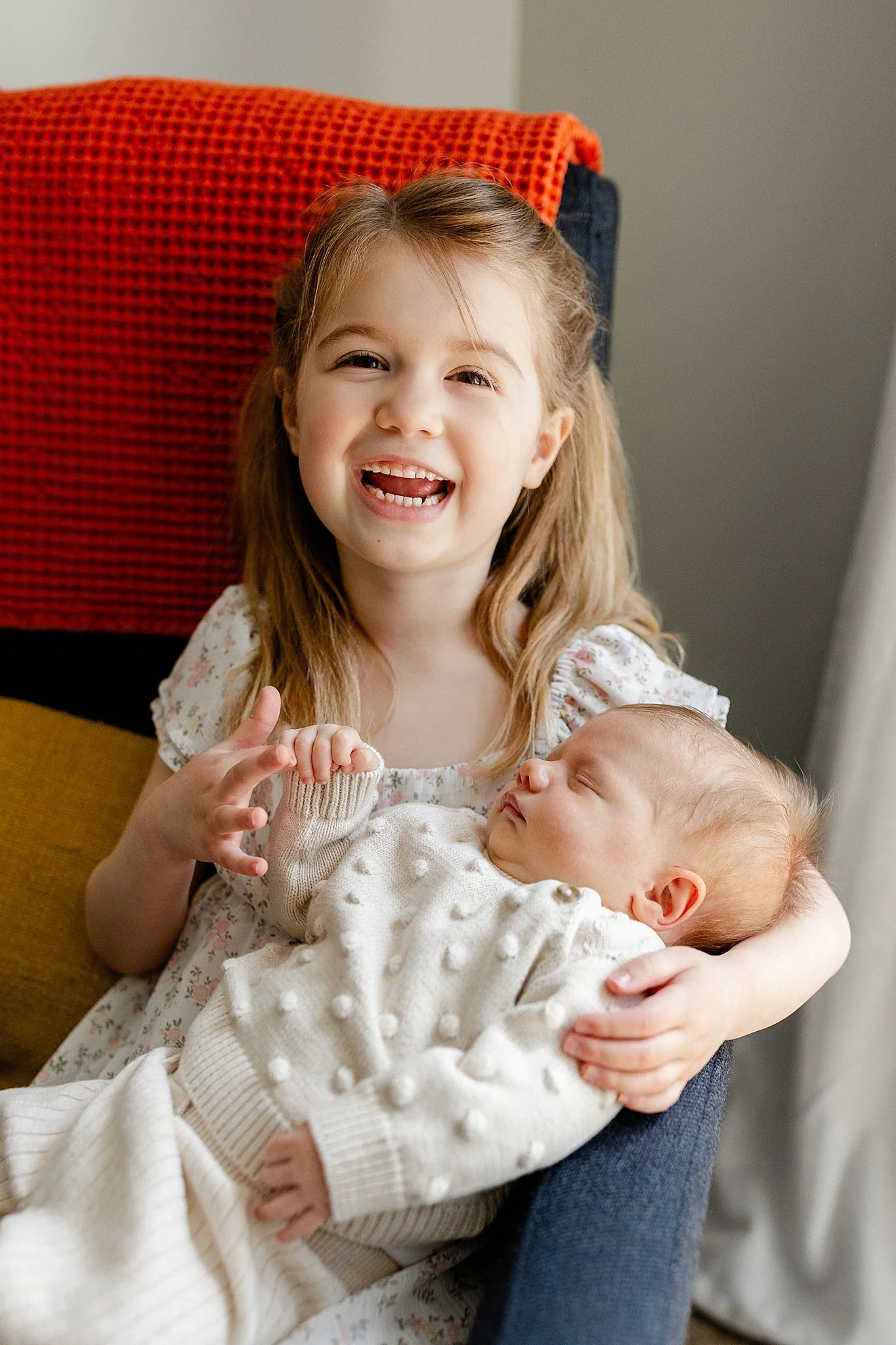 A young girl with blonde hair smiling and holding an sleeping baby wrapped in a white blanket with a lifestyle photographer downtown Naperville.