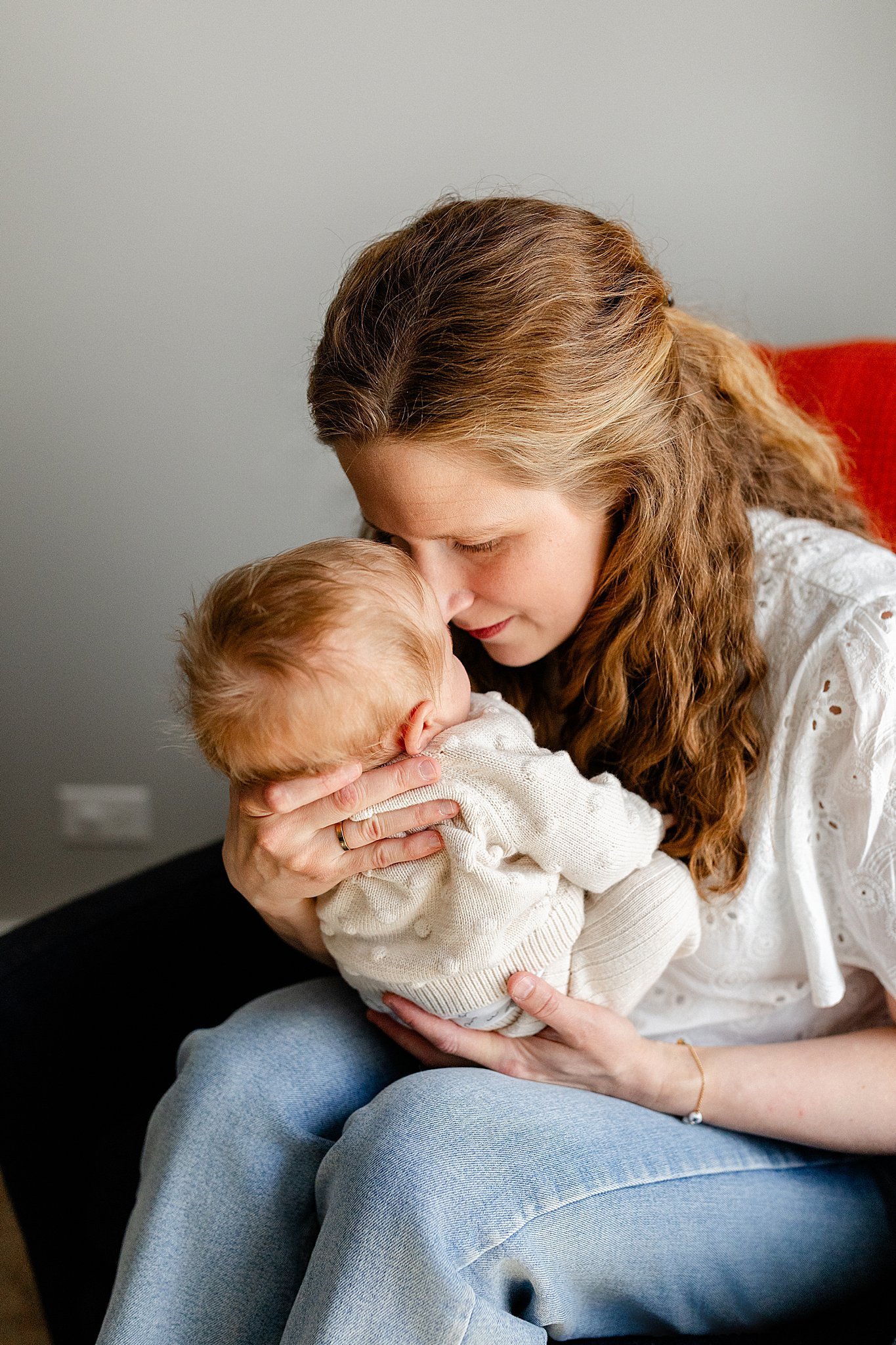 A woman with long, curly brown hair holds a baby with light blond hair and fair skin, pressing their foreheads together in an intimate moment with Ally and B Photography for lifestyle newborn photos.